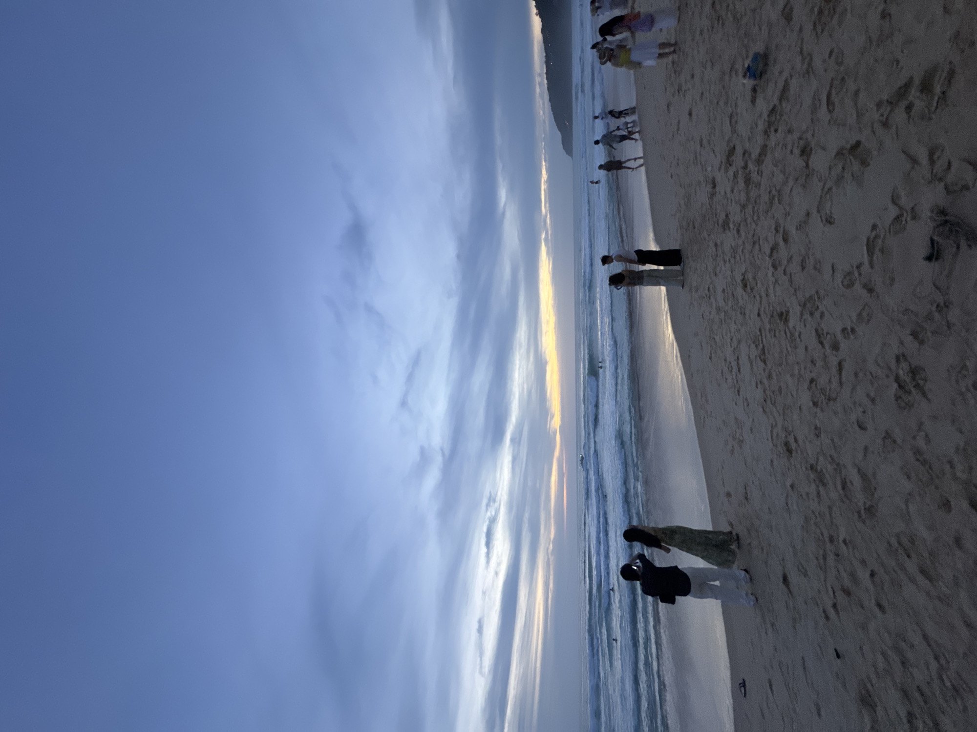The image depicts a beach scene at dusk in Karon, Thailand. A wide expanse of sand occupies the foreground, showing numerous footprints. In the mid-ground, the ocean meets the shore with gentle waves. Several individuals are visible along the coastline and in the water. On the beach, a couple stands near the water's edge, with one person taking a photo. Further down the beach, other groups of people are walking or standing. The sky above is a gradient of deep blue and lighter shades, with streaky clouds illuminated by the setting sun, which is low on the horizon, casting a soft light. A distant headland is visible on the right side of the image. The overall atmosphere is tranquil, suggesting the end of a day at the beach.