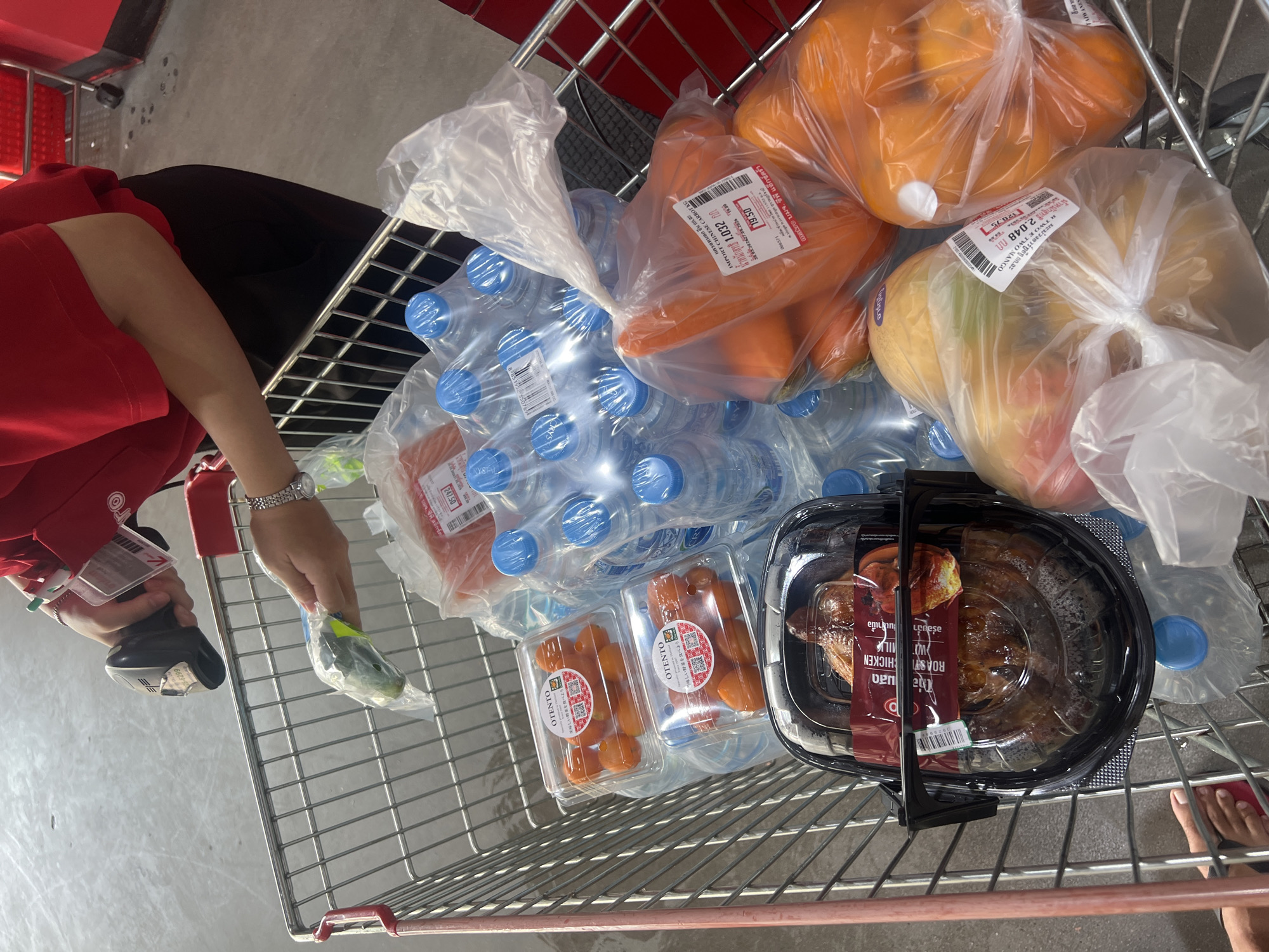 Here is a detailed description of the image:
**Scene Overview:** The image depicts the contents of a shopping cart. There is a person, partially visible, with a red shirt and an ID badge, using a scanner.
**Objects & Subjects:**
* Several packages of bottled water.
* A bag of oranges.
* A bag of carrots.
* A bag of mangoes.
* Two packages of small fruits, labeled "OTENTO".
* A package of roasted chicken.
* A bag of unidentified vegetables
* Scanner
* Shopping cart.
**Environment:** The environment appears to be inside a grocery store. The floor has a simple, light-colored tile or concrete surface.