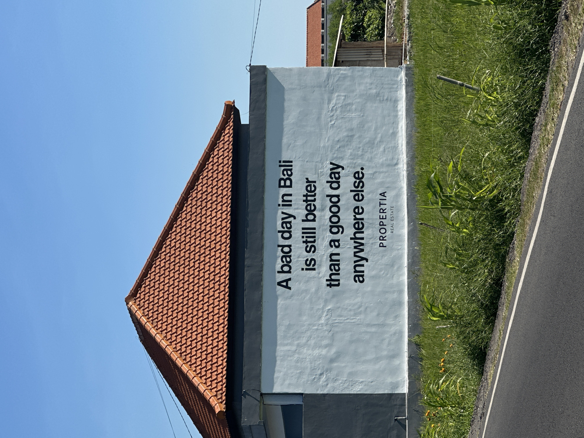 The image shows the side of a building in North Kuta, Indonesia. The building has a terracotta tiled roof. A sign painted on the side of the building reads: "A bad day in Bali is still better than a good day anywhere else." Below the text is the word "PROPERTIA" and the words "REAL ESTATE". Next to the building is overgrown green grass and a road. The sky is blue.
