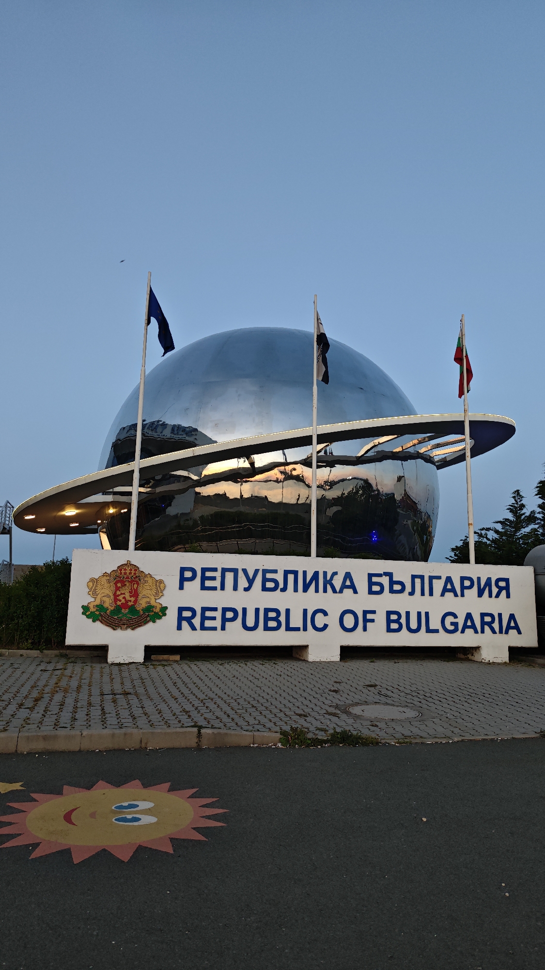 The image shows the entrance to the city of Burgas, Bulgaria, marked by a sign that reads "Република България" and "Republic of Bulgaria" in both Bulgarian and English. The sign also features the coat of arms of Bulgaria. Above the sign is a large, mirrored globe-like structure with a ring around it, reminiscent of the planet Saturn. Three flagpoles stand around the structure, each bearing a flag. The sky is a clear blue, suggesting either early morning or late afternoon. On the ground in front of the sign, there's a painted cartoon sun on the asphalt. The overall scene is peaceful and appears to be a well-maintained area.