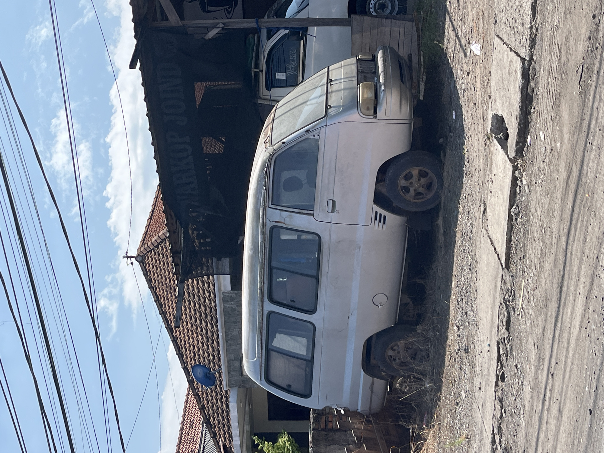 This image captures a scene on a sunny day in Wunut, Indonesia. In the foreground, a worn, silver minivan with visible rust and dirt is parked on a dusty, cracked concrete surface. The minivan has four visible doors on its side, with several windows, some tinted. Its front is partially obscured by a black tarp with white lettering that reads "WARKOP JOIND". Behind the minivan, to the right, a white pickup truck is partially visible. The background shows a house with a red tiled roof and a blue satellite dish. Overhead, a tangle of black electrical wires crisscrosses the sky, which is partly cloudy and blue. The environment appears somewhat unkempt, with dry grass and debris scattered around the vehicles and the ground.