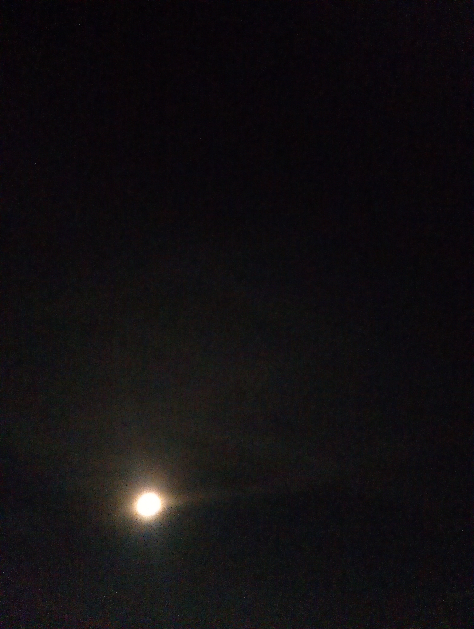 A dimly lit nighttime photograph captures the full moon shining brightly in a dark sky. The moon is positioned in the lower left quadrant of the image, emitting a soft glow that slightly illuminates the surrounding dark expanse. The sky appears mostly devoid of clouds, with a few faint speckles of light visible, potentially distant stars or noise in the image. The overall impression is one of serene darkness with the moon as the sole prominent celestial body. The photograph does not provide any identifiable landmarks or context that would allow for pinpointing a specific location within Vitsebsk, Belarus.