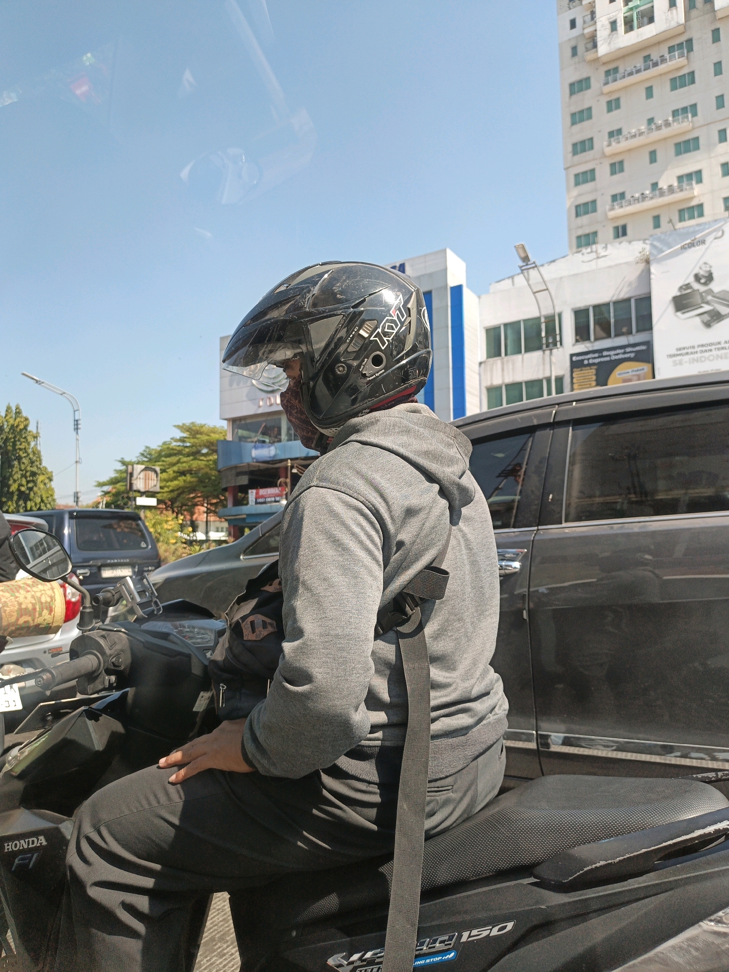 A person wearing a black full-face helmet and a grey hooded sweatshirt is sitting on a motorcycle in what appears to be a city environment. The person has a patterned mask covering their lower face. The motorcycle is black, with "HONDA FI" visible on the side. The person's left hand is resting on their thigh, and a black strap from a bag or backpack is visible over their shoulder and down their back.
In the background, there is a dark-colored car, a blue SUV, and a tall, modern building with many windows and balconies. Another building, shorter and white with blue accents, is to the left of the taller building. There are trees and streetlights visible. The sky is clear and blue. The scene suggests traffic, possibly at an intersection or in a busy street. The presence of specific building designs and the "HONDA FI" branding on the motorcycle, along with common vehicle types, point to an urban setting in Indonesia, consistent with the location of Surakarta.