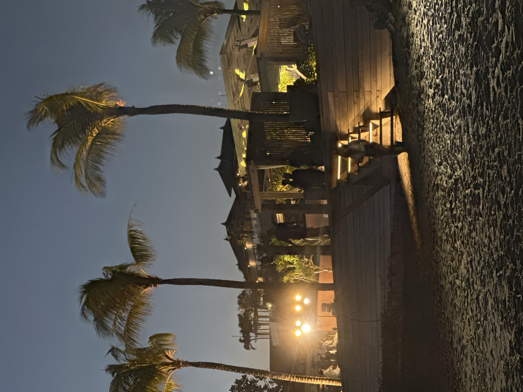 This nighttime photograph, taken in North Kuta, Indonesia, depicts an outdoor resort or hotel scene illuminated by artificial lights against a dark blue evening sky.
On the left side, several tall palm trees with vibrant fronds are silhouetted, signifying a tropical setting. Across the middle ground, multiple multi-story buildings feature distinct architecture, likely of Indonesian influence, with dark wood construction and pointed roofs. These structures are warmly lit, highlighting their details and balconies.
Wooden walkways and stairs wind through the area, with numerous lights integrated into the steps, providing guidance and ambiance. To the right, a large, ruggedly textured wall or ground surface, possibly dark stone or concrete, is illuminated to emphasize its rough pattern.
Further in the background, an area is brightly lit by what appear to be stage lights or powerful floodlights. Here, several individuals are visible, mostly in silhouette or dimly lit, suggesting a social gathering, an outdoor dinner, or a performance. The overall scene evokes a luxurious and relaxed tropical evening, bustling with subtle activity. No discernible text is visible within the image.