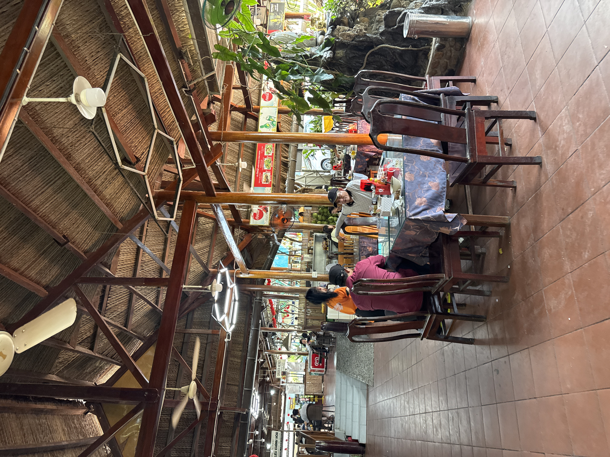 The image depicts the interior of a large, open-air restaurant with a thatched roof supported by wooden beams and pillars. The floor is tiled with reddish-brown square tiles. In the foreground, a round table is set with a floral patterned tablecloth, plates, bowls, and condiments. Four wooden chairs surround the table. Three people are seated at the table, one facing away from the camera and two facing towards it. A fourth person, wearing an orange shirt, stands behind one of the seated individuals.
Further into the restaurant, other tables and chairs are visible, with some patrons seated. The background shows more of the restaurant's interior, including signage with Vietnamese text and images of food. There are also decorative elements such as plants and a rock formation with a statue. Multiple ceiling fans are mounted on the wooden beams. The overall atmosphere suggests a casual dining environment. The location is identified as Nam Khanh Vinh, Vietnam.