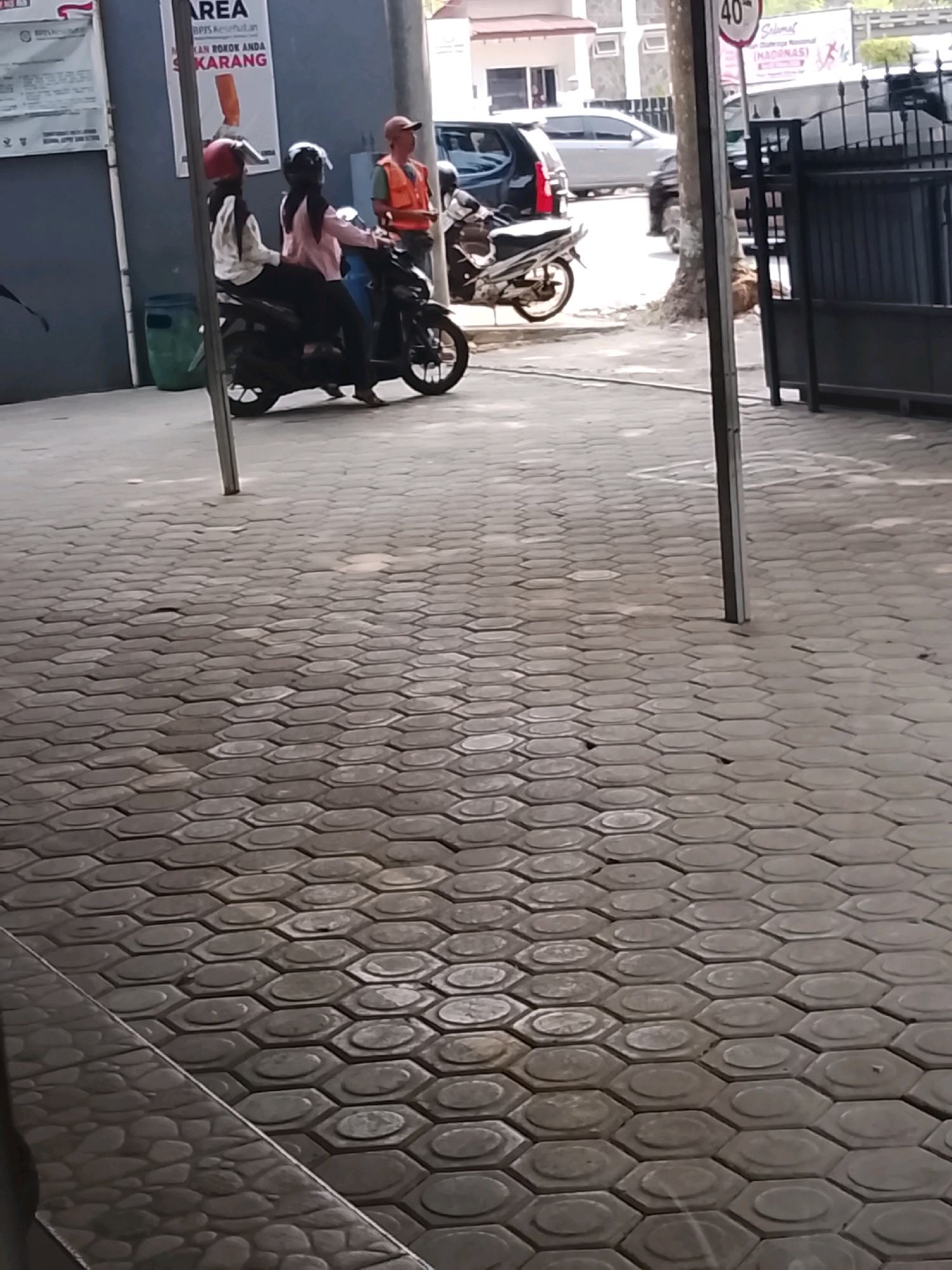 The image depicts an outdoor scene, likely on a paved walkway in the city of Slawi, Indonesia. In the center, two individuals wearing helmets are seated on a black motorcycle. The person in the front, on the left, wears a red helmet and a white jacket with black accents. The person behind them, on the right, wears a black helmet and a light pink jacket. To the right of the motorcycle, a person wearing an orange vest and a dark baseball cap stands near a fence. Behind the motorcycle, a silver car and a white vehicle are visible. A tall, dark metal fence with vertical bars is prominent on the right side of the image. The ground is paved with hexagonal interlocking bricks. A partially visible trash bin is located to the left of the motorcycle. A vertical metal pole stands in the foreground, obscuring part of the scene. Signs are visible on a building wall in the background.
