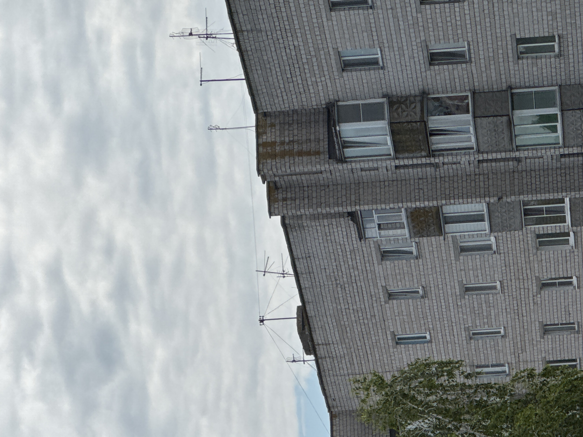 This is a photograph of the upper section of a grey brick apartment building, likely in a residential area. The building has multiple windows and balconies. Several television antennas are visible on the rooftop. A tree with green foliage is partially visible on the left side of the frame. The sky is overcast with grey clouds. The scene appears to be captured during daylight hours. The location is specified as Yarominski Sel'ski Sovet, Belarus.