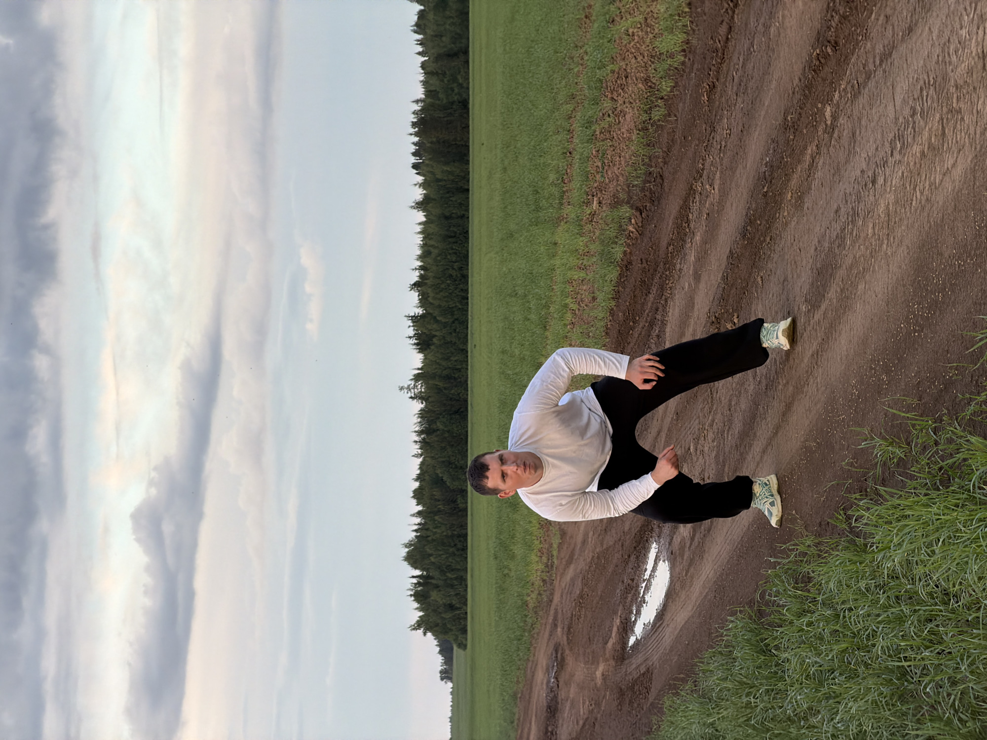A man in a white long-sleeve shirt and black pants stands on a muddy path in Фоки, Russia. He is bent over slightly, with one hand on the ground and the other bent near his hip. The path is flanked by a green field on one side and a forest line on the other. The sky is cloudy.