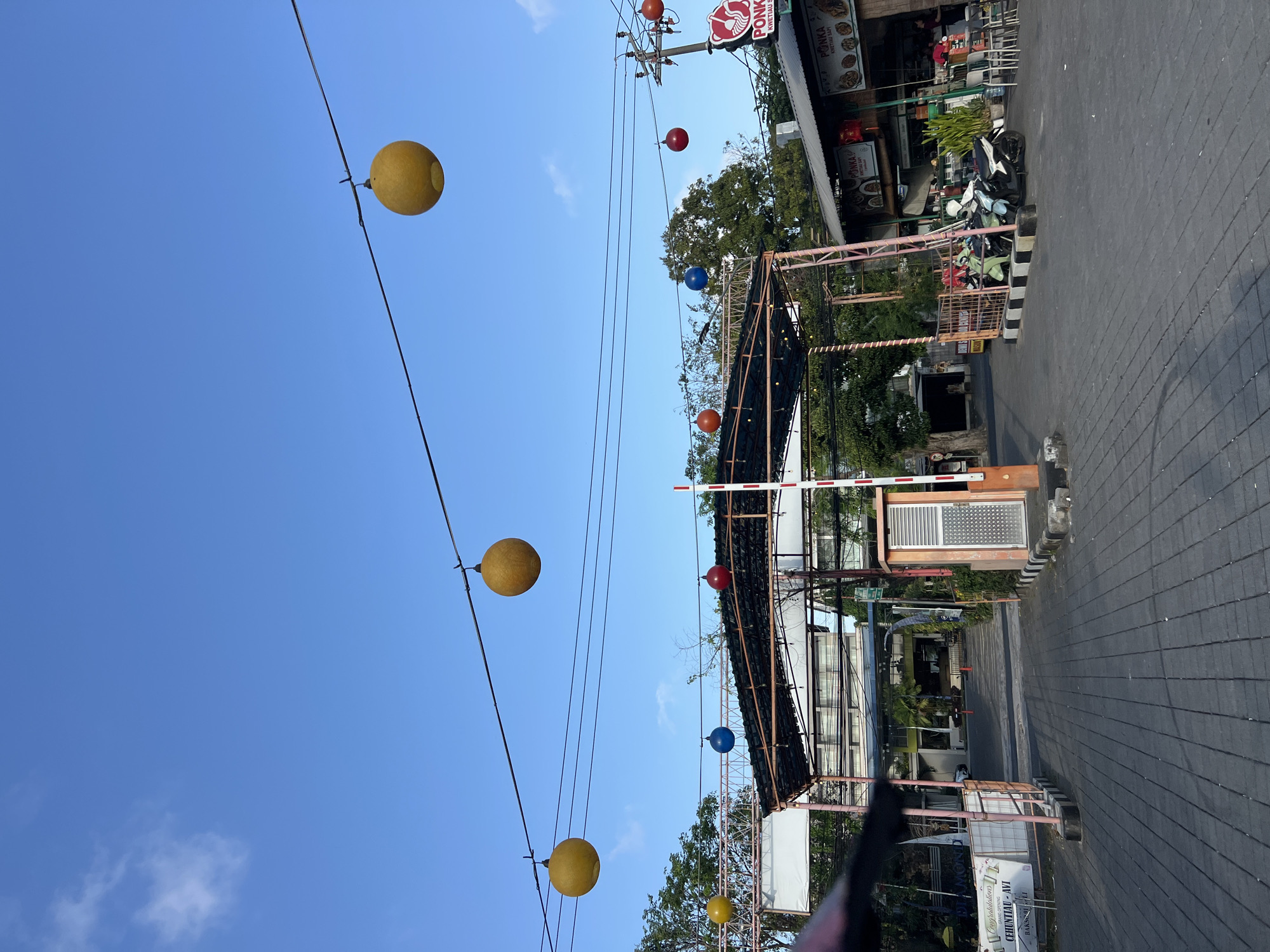 In Kuta, Indonesia, an outdoor scene unfolds under a clear blue sky with scattered clouds. The foreground features a gray brick-paved driveway leading towards a covered structure with pink and white striped posts. Overhead, a series of black wires are strung, supporting several brightly colored, spherical lanterns: yellow ones are prominent, along with a blue and a red one. To the right, a white and red striped barrier gate stands next to a gray and orange metal booth. Beyond the gate, a row of low-rise buildings houses various food establishments, indicated by signage such as "PONKA KWETIAU SAPI." People are visible in these areas, some seated at tables outside the restaurants and others on motorcycles. The background reveals a building with multiple stories, surrounded by lush green trees. The overall impression is that of a lively, well-lit commercial area, possibly a food court or market.