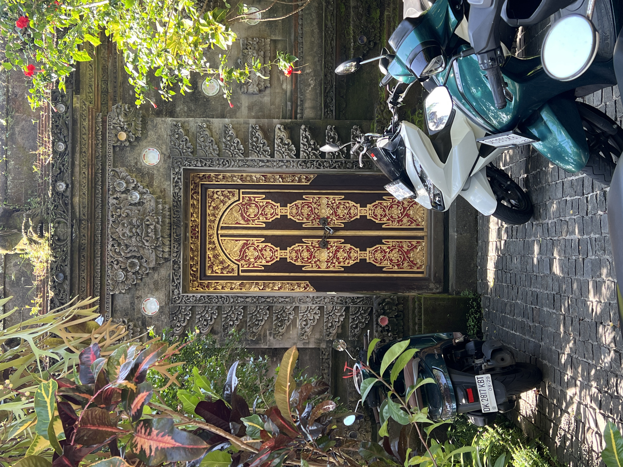 The image depicts a street scene in Petulu, Indonesia. An ornately carved, brown and gold wooden door is centered in the frame, set into a grey stone wall decorated with carvings and round accents. Vegetation frames the door from below, including red and green plants. Two scooters are parked on the right side of the image. One scooter is dark green and the other is white and green. The license plate DK 2817 KBT is visible on the green scooter. The ground appears to be paved with grey stone blocks. The image is taken during the day, and the lighting suggests a sunny day with some shadows. Some plants and tree branches are visible above the stone wall and door.