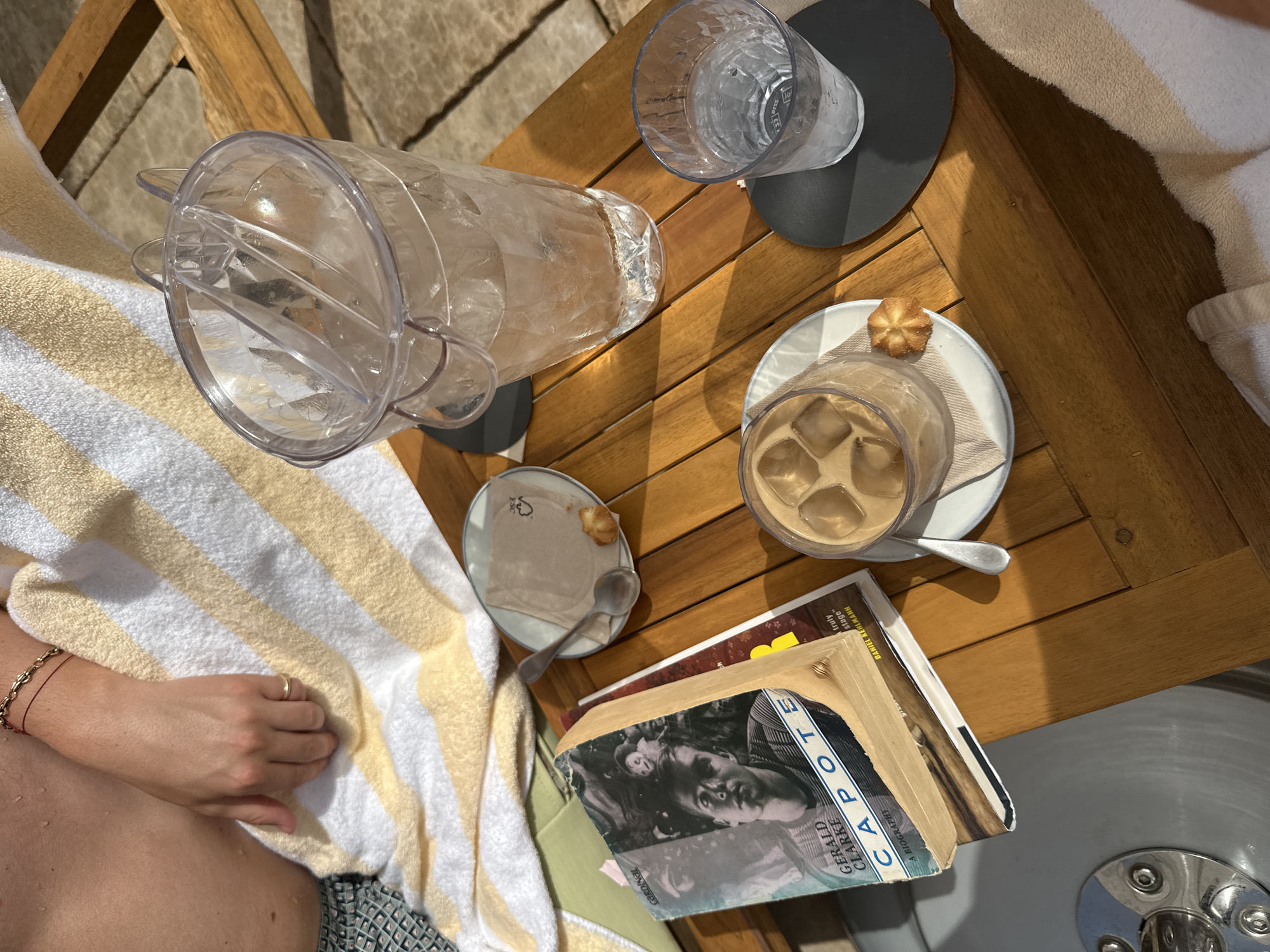 The image shows a close-up overhead view of a wooden table next to a body of water, likely a swimming pool. On the table are several items, including a book titled "Capote" by Gerald Clarke, a glass of iced coffee with ice cubes, a glass of water with ice cubes, an ice bucket filled with ice and water, a small plate with a napkin and a cookie, and a spoon. A person's arm and torso are visible on the left side of the image, draped with a striped towel, suggesting they are relaxing in a lounge chair. The overall setting appears to be an outdoor recreational area.