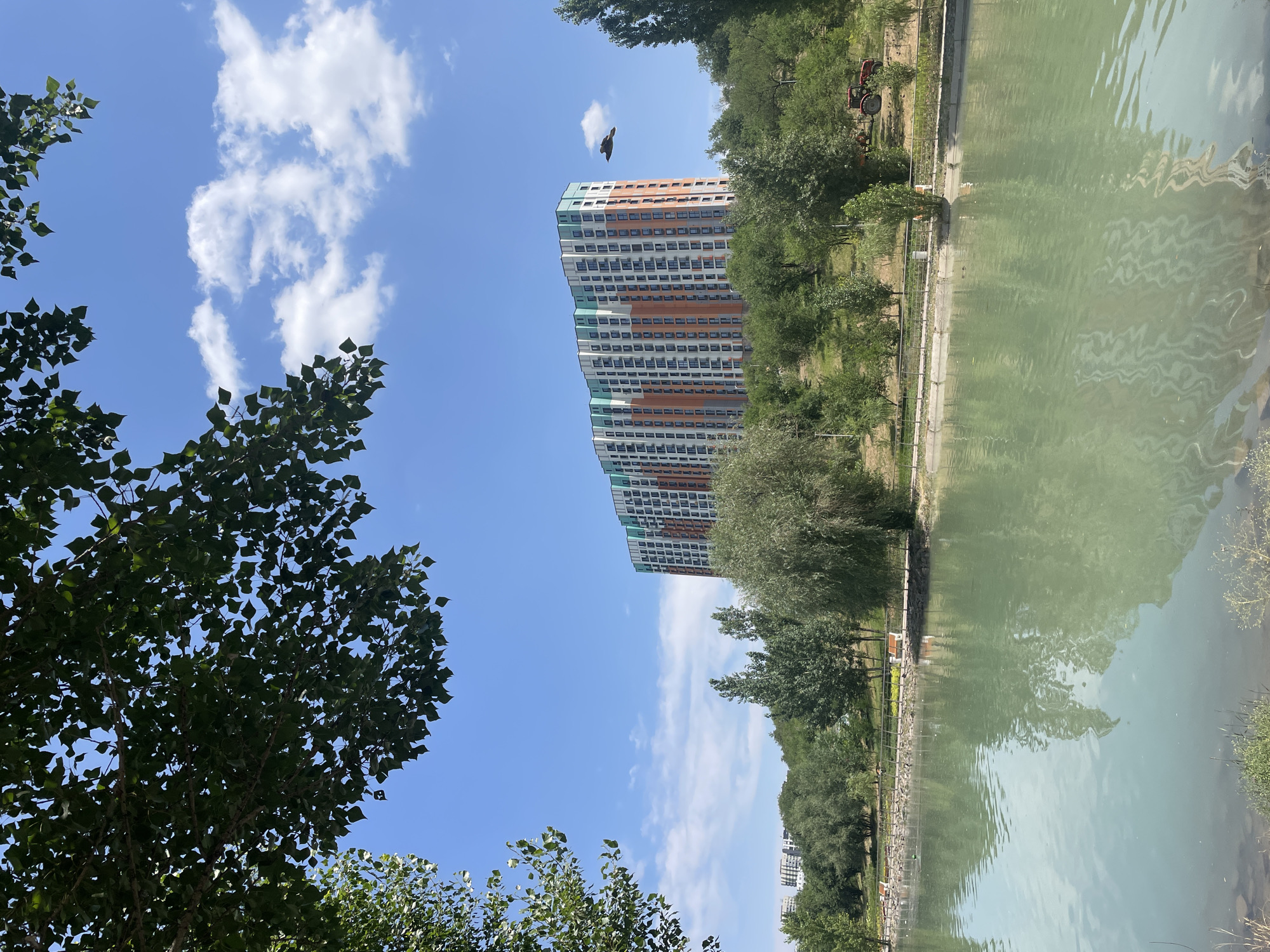 This media file showcases an outdoor scene with a large, colorful apartment building as a central subject. The building features a facade of white, orange, and teal colored panels, with numerous windows visible. In the foreground, lush green trees with dark leaves frame the left side of the image, obscuring part of the sky. A bright blue sky with scattered white clouds dominates the upper portion of the frame.
A bird is captured mid-flight in the sky, appearing as a dark silhouette against the clouds. To the right of the apartment building, a body of water, likely a pond or canal, reflects the sky and surrounding greenery, creating a shimmering effect on its surface. Along the bank of the water, a fence and some vegetation are visible. Further to the right, a red tractor is parked amongst trees. The overall impression is a serene, daytime scene with a mix of urban and natural elements. The presence of the tractor suggests some sort of activity or maintenance might be occurring in the area, but no people are directly visible. The weather appears sunny and clear, typical for a pleasant day. The location context provided is Almaty, Kazakhstan. No visible text is present in the image.