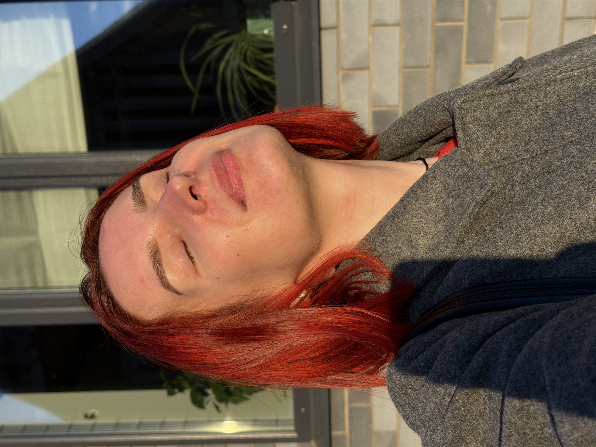 A medium, angled, outdoor shot captures a person with bright red hair, eyes closed, facing upwards towards the sun. The person is wearing a gray woolen coat. The background consists of a window reflecting the sky, a brick wall, and some foliage, possibly a houseplant. The image was taken in a sunny location, based on the light falling on the person's face and coat. There is not sufficient information to verify the location in the city of Chaikovsky, Russia, based on the visual data.