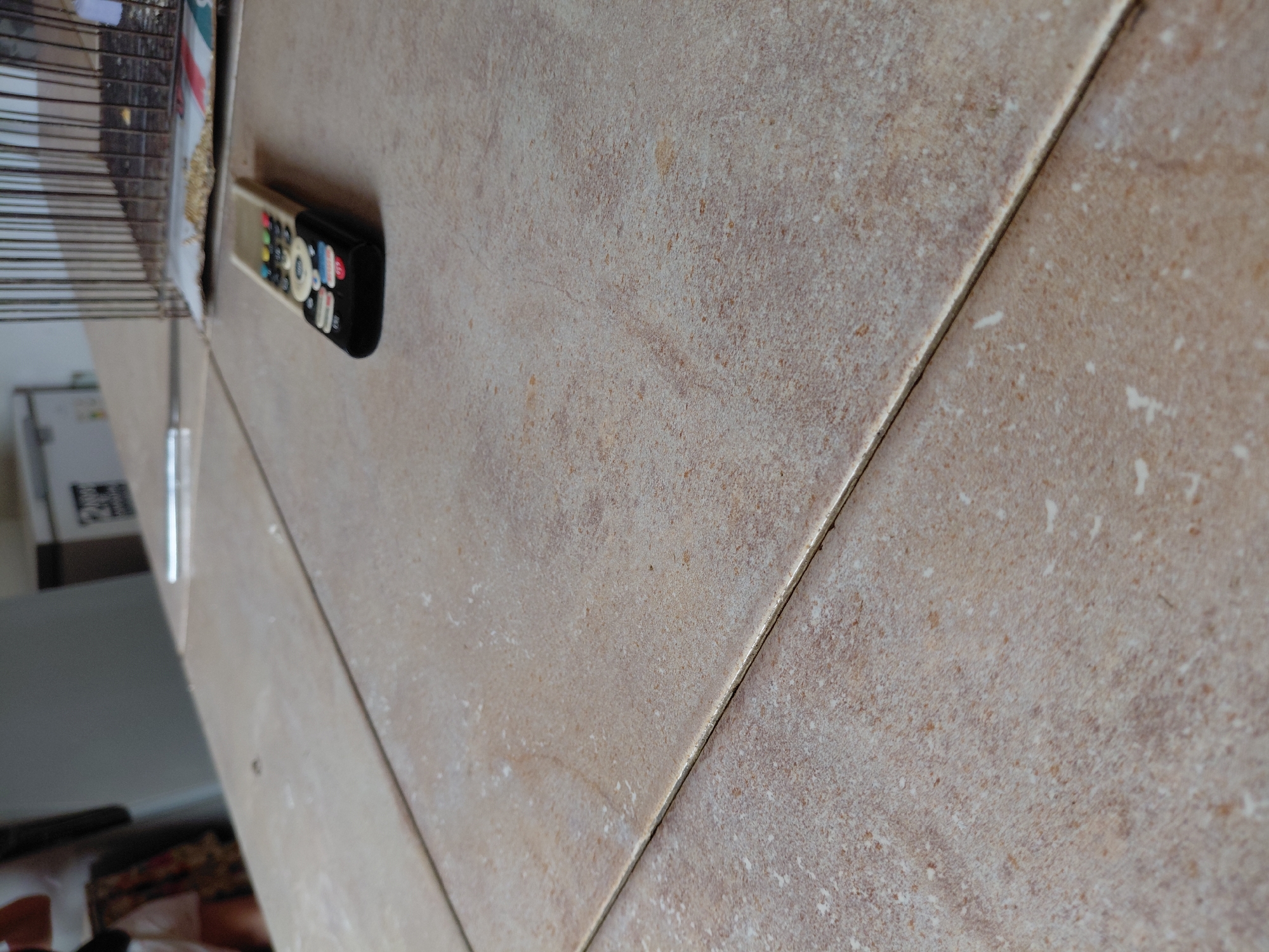 This media file, taken in Santa Ana, Venezuela, captures an indoor scene dominated by a light brown or beige tiled surface. The tiles feature a speckled, somewhat rough texture, resembling ceramic or natural stone, and are clearly separated by dark grout lines.
In the mid-ground, a black remote control is resting on the tiled surface. It has a beige upper half and black bottom half, with various colorful buttons, including a prominent red Netflix button and other colored app icons that are difficult to fully discern due to the angle and focus.
To the left, a blurry object resembling a metal cage or wire rack is partially visible, containing granular material, possibly birdseed. In the distant, blurry background, a white appliance, likely a refrigerator, can be seen, featuring a small, unreadable black and white label or QR code. The lighting suggests a daytime setting, possibly with indirect natural light or consistent indoor illumination. No activity is taking place, and no people are visible in the static shot.