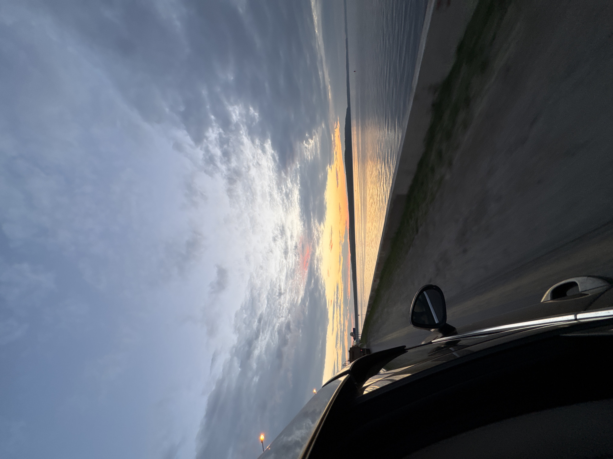 This image captures a view from the passenger side of a moving car at dusk, looking out towards a body of water. The sky is a blend of muted blues and grays with streaks of orange and pink from the setting sun near the horizon. Clouds are present throughout the sky, some thin and wispy, others darker and more solid. The water reflects the colors of the sky, creating a calm and expansive surface. To the right of the water is a concrete embankment. Adjacent to the embankment is a paved road, with a narrow strip of grass and vegetation between the road and the embankment. The car's side mirror and the edge of the car's body are visible in the foreground on the left. The road appears to be a straight stretch with some distant traffic lights and a truck visible further down the road. This scene is likely captured along the Kama River embankment in the city of Chaikovsky, Russia, known for its scenic waterfront views.