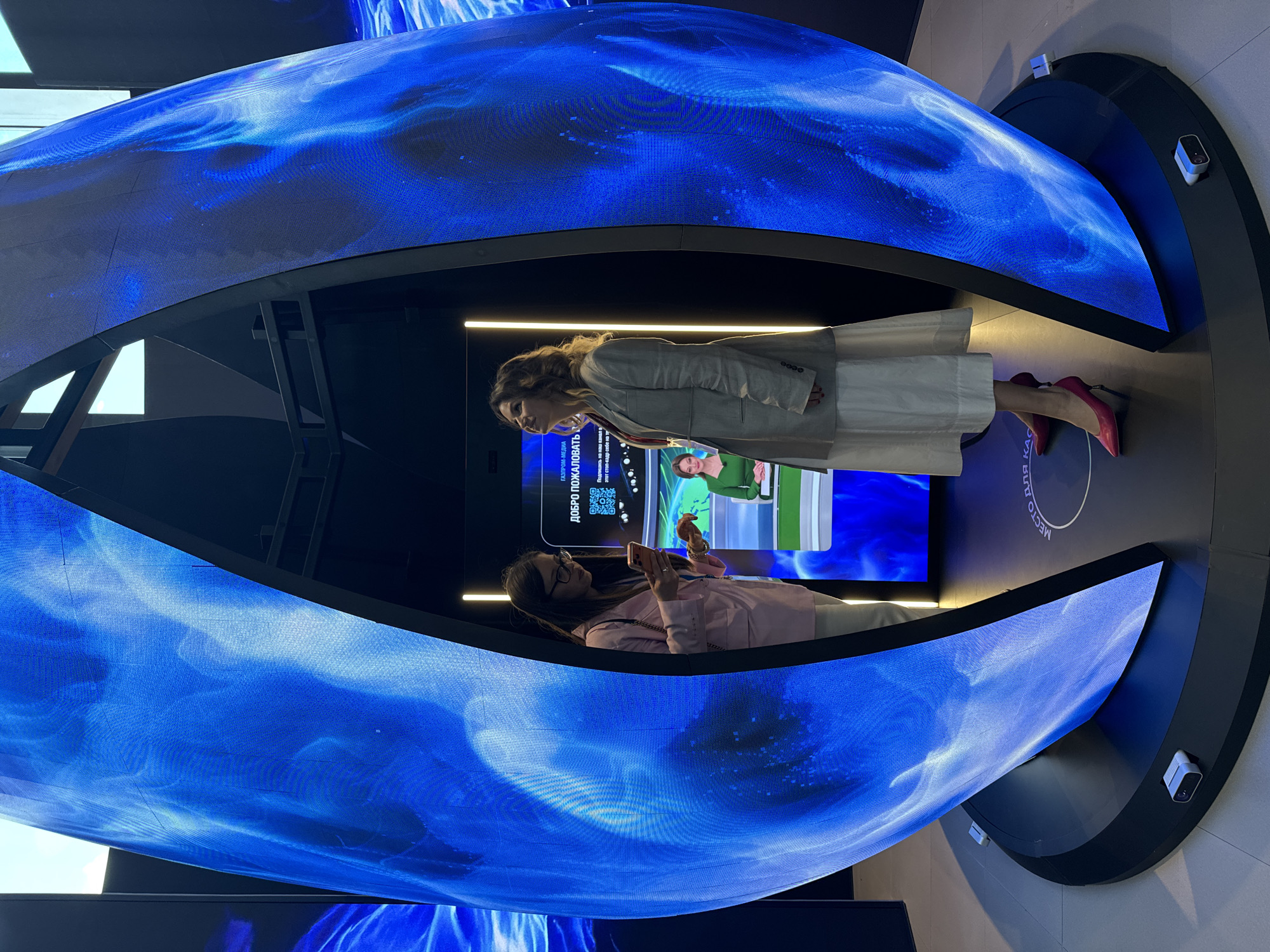 An indoor, modern exhibition setting in Shushary, Russia, features a large, curved LED display creating a tunnel-like, immersive environment. The display showcases dynamic blue, abstract patterns reminiscent of water or cosmic scenes.
Two women are present, observing the digital installations. One woman, dressed in a light-colored blazer and a white skirt with red high heels, stands facing the main display, engrossed in its content. Her back is slightly turned towards the camera, and her reflection is visible on the inner surface of the curved screen. Directly opposite her, a second woman, wearing glasses and a pink top, is holding a smartphone, seemingly capturing an image or video of the screens. She also appears engaged with the visual content.
In the background, behind the first woman, a flat screen shows a news-like broadcast featuring a woman presenter. The platform they are standing on has a circular design element. Security cameras or sensors are mounted on the wall near the curved display.
Visible text on the screens includes Russian phrases: "ГАЗПРОМ-МЕДИА" (GAZPROM-MEDIA), "ДОБРО ПОЖАЛОВАТЬ" (WELCOME), and partially visible phrases such as "этот стоп-кадр себе на тр" (this freeze-frame for yourself on tr...) and "ДЛЯ КАС" (FOR CASH / FOR KAS...). A QR code is also displayed on the flat screen. The scene suggests an interactive media or technology showcase designed to captivate visitors.