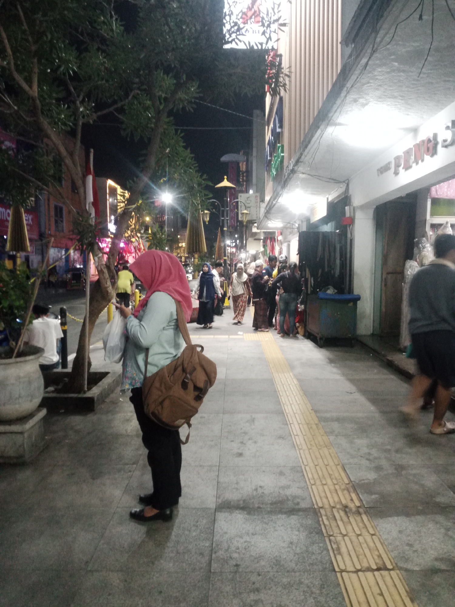 This image captures a nighttime scene on a bustling pedestrian walkway in the city of Tasikmalaya, Indonesia. The walkway is paved with gray stone tiles, with a distinct yellow tactile paving strip running alongside. To the right, a building with a white facade and ribbed siding is visible, displaying the signage "PENG S" and another illegible word in red. Storefronts with closed doors and displays of merchandise are present. The left side of the walkway features trees with dense foliage, a large potted plant, and several lamp posts, one of which is decorated with ornamental umbrellas.
In the foreground, a woman wearing a light blue long-sleeved top, black pants, and a pink hijab is seen from the side, looking down at her phone. She carries a brown backpack over her right shoulder and holds a plastic bag in her left hand. Further down the walkway, a group of people are walking and standing, some in traditional attire. A person in a yellow shirt is visible in the background, and another individual in a gray sweater and dark shorts walks away from the camera on the right side of the image. The scene is illuminated by streetlights and the lights from the shops, creating a moderately lit urban environment.