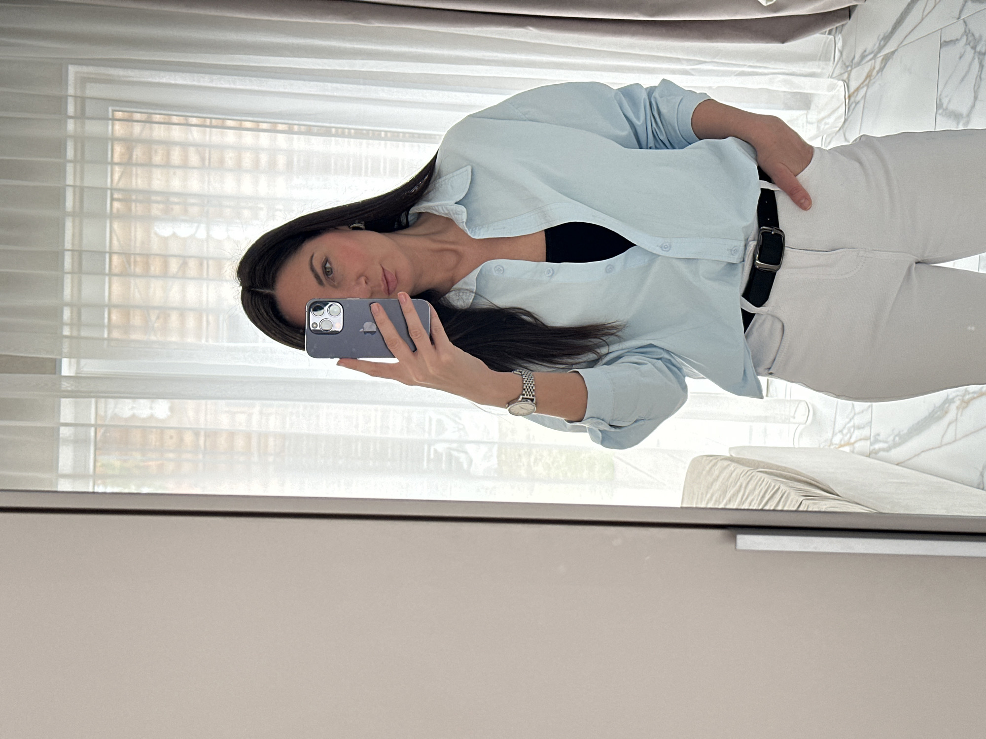 An indoor, mirror selfie features a woman captured from the waist up, dressed in contemporary attire. The image is presented as if correctly oriented, with the woman facing slightly leftward, looking directly into the camera reflected in the mirror. She has long, dark hair, which falls over her left shoulder and chest. Her skin is fair, and she has dark, defined eyebrows and dark eyes, with a neutral expression.
She is wearing a light blue, long-sleeved button-up shirt, unbuttoned to reveal a black top underneath. Her lower body is clad in white trousers, secured by a black belt with a rectangular silver buckle. A silver-toned wristwatch with a metal band is visible on her left wrist.
In her right hand, she holds a grey smartphone with a prominent multi-lens camera system, indicating she is taking the selfie. The background, reflected in the mirror, shows natural light filtering through a large window covered with white blinds or sheer curtains, suggesting daytime. To the right of the woman, a light-colored sofa or armchair is partially visible, alongside a white and grey marble-patterned surface, possibly a wall or floor. The lower part of the mirror's reflection reveals a section of a light brown cabinet or furniture piece with a silver handle. The setting appears to be a modern residential interior in Chaikovsky, Russia.