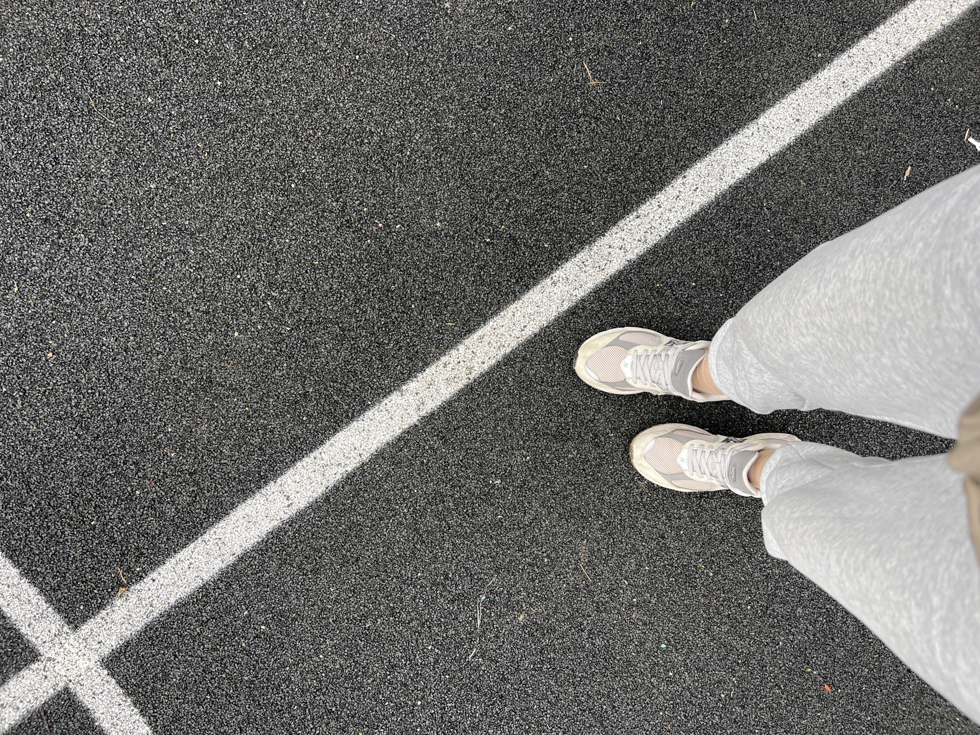 An overhead shot captures a person's lower legs and feet standing on a dark, granular, rubberized surface, likely a sports court or running track, in Khimki, Russia. The person is wearing light grey sweatpants and light-colored athletic shoes, possibly sneakers, with visible laces. Bright sunlight casts sharp shadows of white lines painted on the dark ground. One prominent white line runs diagonally from the bottom-left to the top-right of the frame, intersecting with another white line in the bottom-left quadrant, forming a corner or intersection marking. The ground surface appears textured, composed of many small, dark grey or black specks. The strong shadows indicate a clear, sunny daytime. No discernible activity beyond standing is depicted, and no legible text is visible on the clothing, shoes, or ground markings.