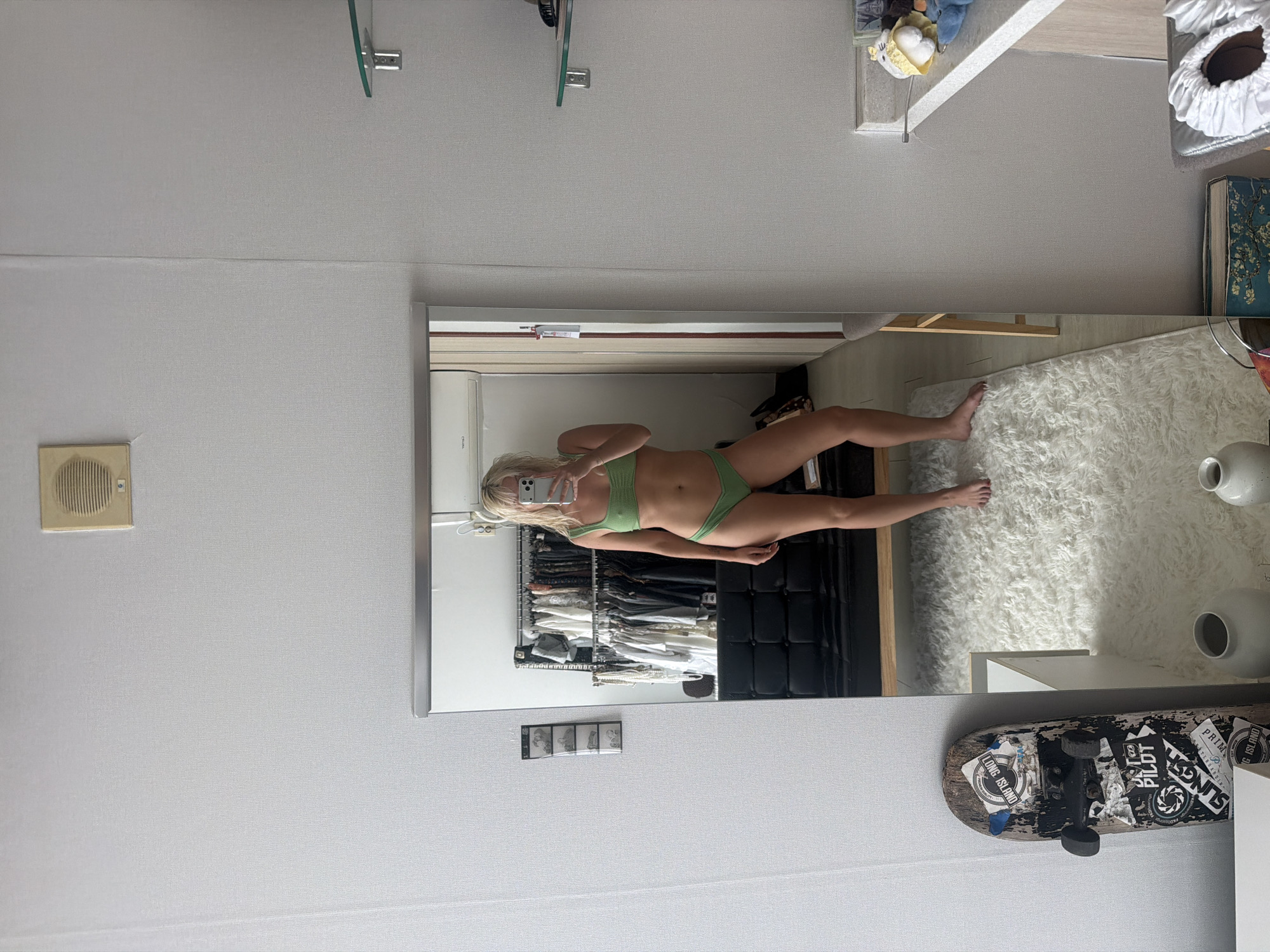 The image is an indoor shot depicting a woman reflected in a mirror. The woman, standing in what appears to be a closet or dressing area, is holding a smartphone and taking a selfie. She is wearing a green bikini. The closet behind her is filled with clothes hanging on racks. The mirror also reflects a portion of a room with a white rug and various objects including decorative vases and a skateboard with stickers. The environment has a contemporary, domestic feel. The image provides no evidence to determine the location is the city of Seoul, South Korea.