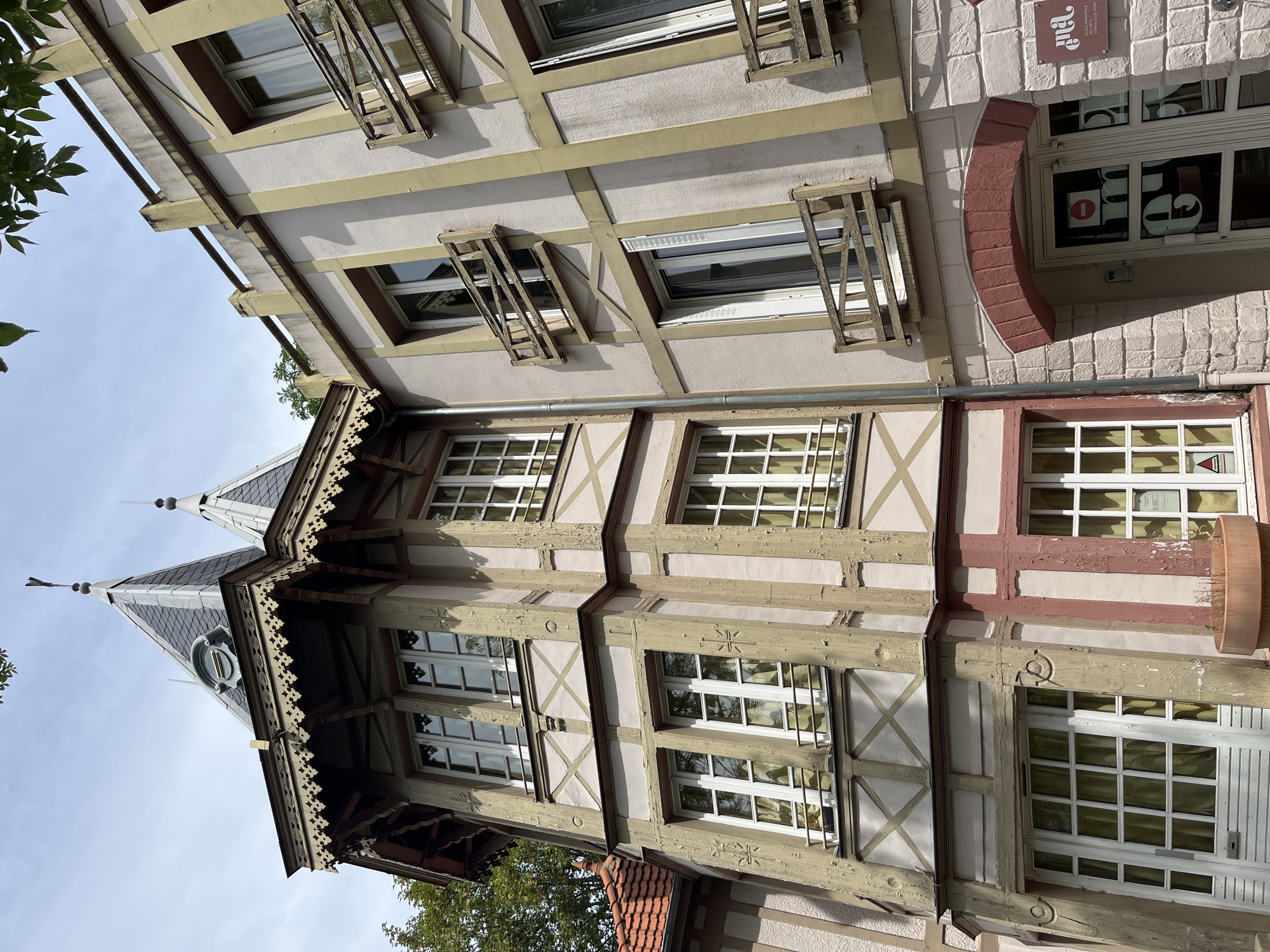 This is a low-angle, eye-level, full shot of the exterior of a building in Joinville-le-Pont, France. The building features a mix of architectural styles, including a prominent tower with a pointed roof, decorative trim, and visible timber framing. The walls are painted a pale pink and cream with yellow trim. Several windows, many with multiple panes, are visible, some with simple wooden balconies. The lower part of the building has a brick facade and an arched entranceway with a sign that reads "ÉCOLE MUNICIPALE DES ARTS." The sky is partly cloudy.