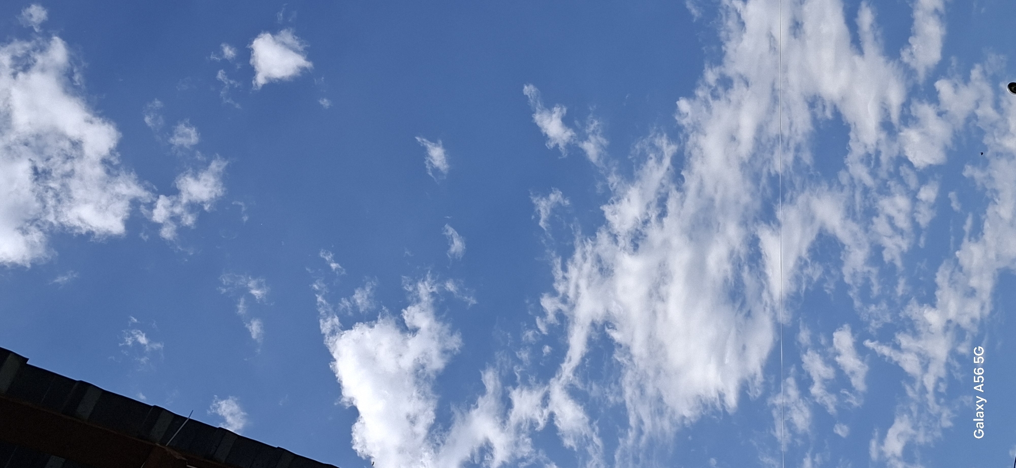 The image is a vertical, low-angle shot of a bright blue sky filled with scattered white clouds. In the upper left corner, a dark, textured surface, possibly a roof overhang or awning, partially obstructs the view. A thin, light-colored horizontal line, likely a wire, stretches across the middle portion of the sky. The bottom left corner of the image displays the text "Galaxy A56 5G." The scene is outdoors and appears to be taken in daylight. There are no people or discernible landmarks that would indicate a specific location in Tbilisi, Georgia. The focus is entirely on the sky and clouds.