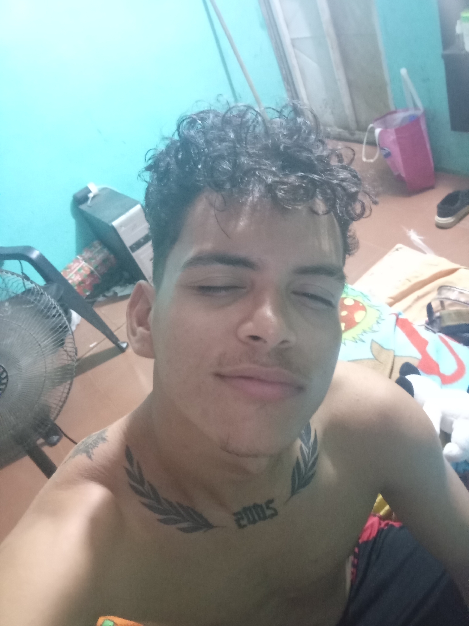 A close-up selfie of a shirtless young man with curly hair, eyes closed. He has tattoos on his chest, one appearing to be a laurel branch on his left shoulder and "2005" centered on his chest above his sternum. An electric fan is on his left. In the background, there is a turquoise wall, and various household items including a pink bag, a pair of shoes, and a box wrapped in decorative paper are visible. The location and city, Altagracia, Venezuela, are not discernible from the image itself.