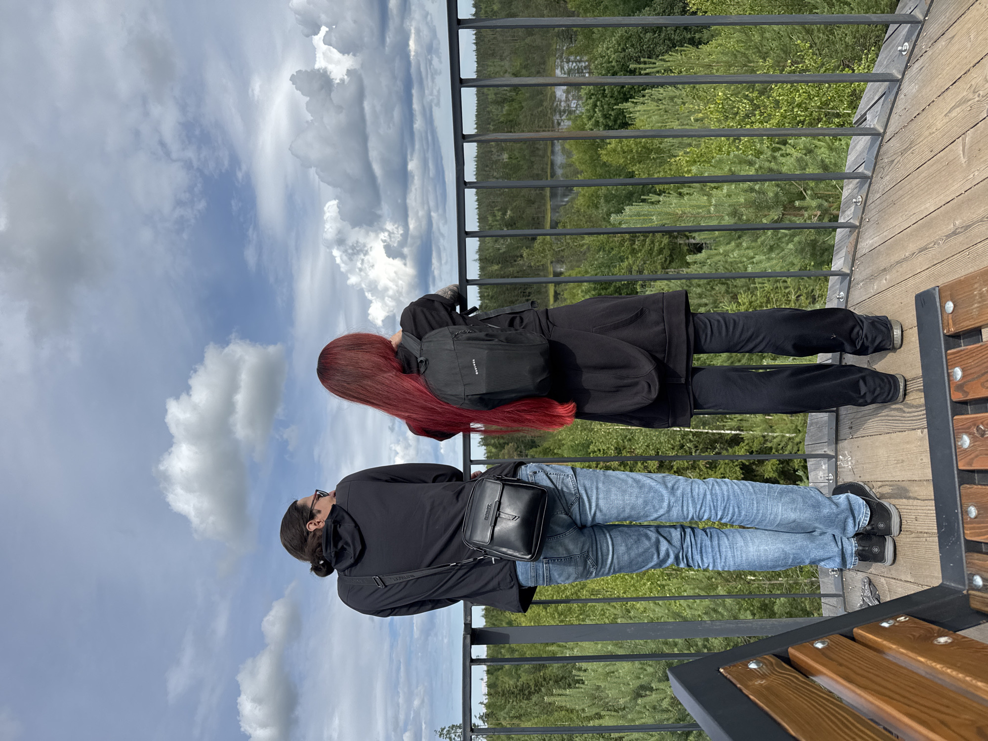 Two individuals, one with dark hair pulled back in a bun and wearing a black hooded jacket, blue jeans, and a black crossbody bag, and another with long, red hair wearing a black hooded jacket, black pants, and a black backpack, stand on a wooden platform overlooking a dense forest. A black metal railing separates the platform from the forest, which slopes down to a body of water. The sky above is partly cloudy, with large, white cumulus clouds against a blue background. A wooden bench is partially visible in the foreground. The scene is likely an observation deck in or near the city of Peski, Russia, offering a panoramic view of the surrounding natural landscape.
