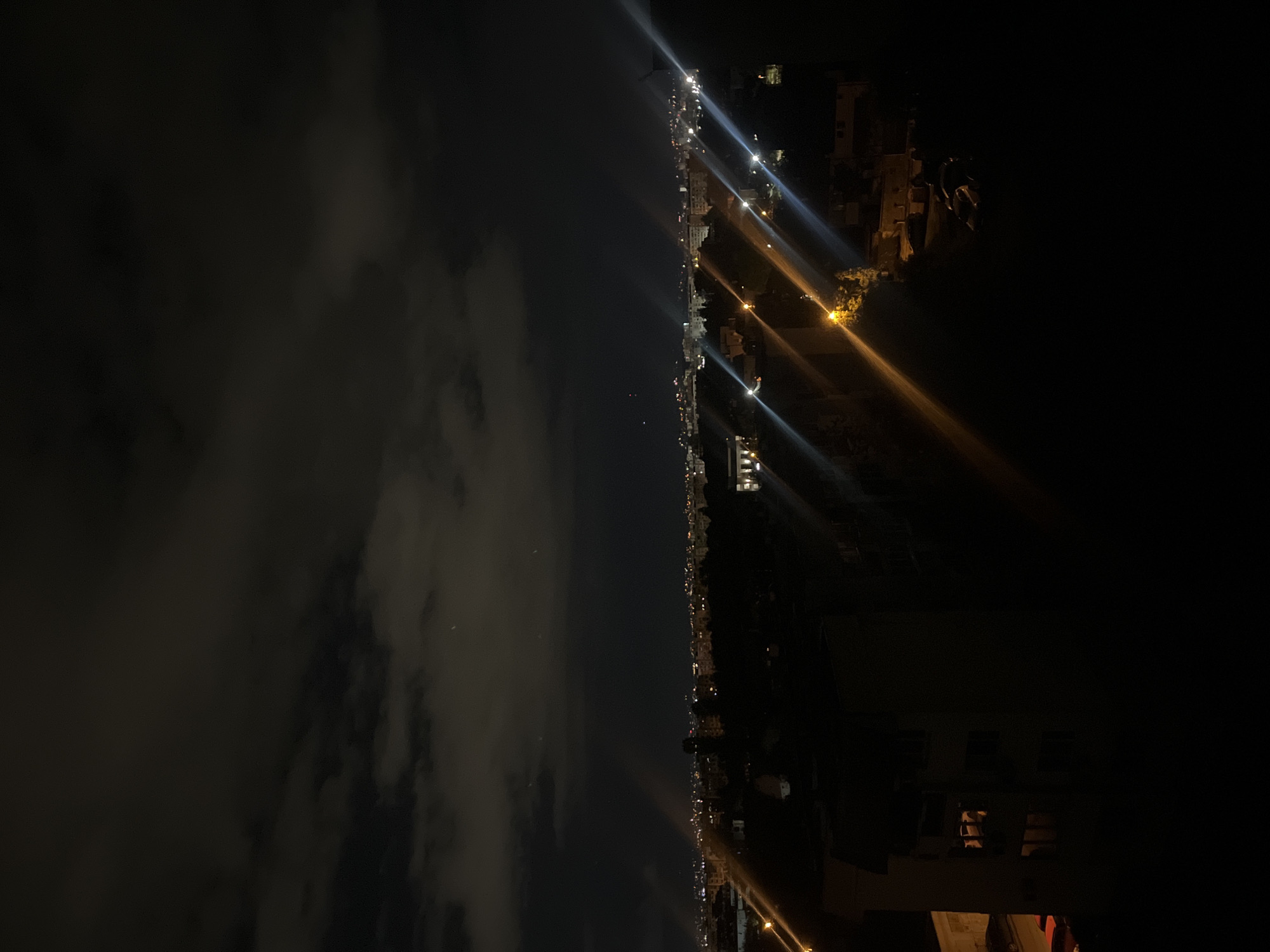 The image depicts a nighttime scene of a city skyline, likely in Sofia, Bulgaria, based on architectural styles and urban layout. The left side of the image is dominated by a dark, cloudy sky. The right side features the city, with scattered lights indicating buildings and streets. Several bright light sources create lens flares. A dark area occupies the lower right corner.