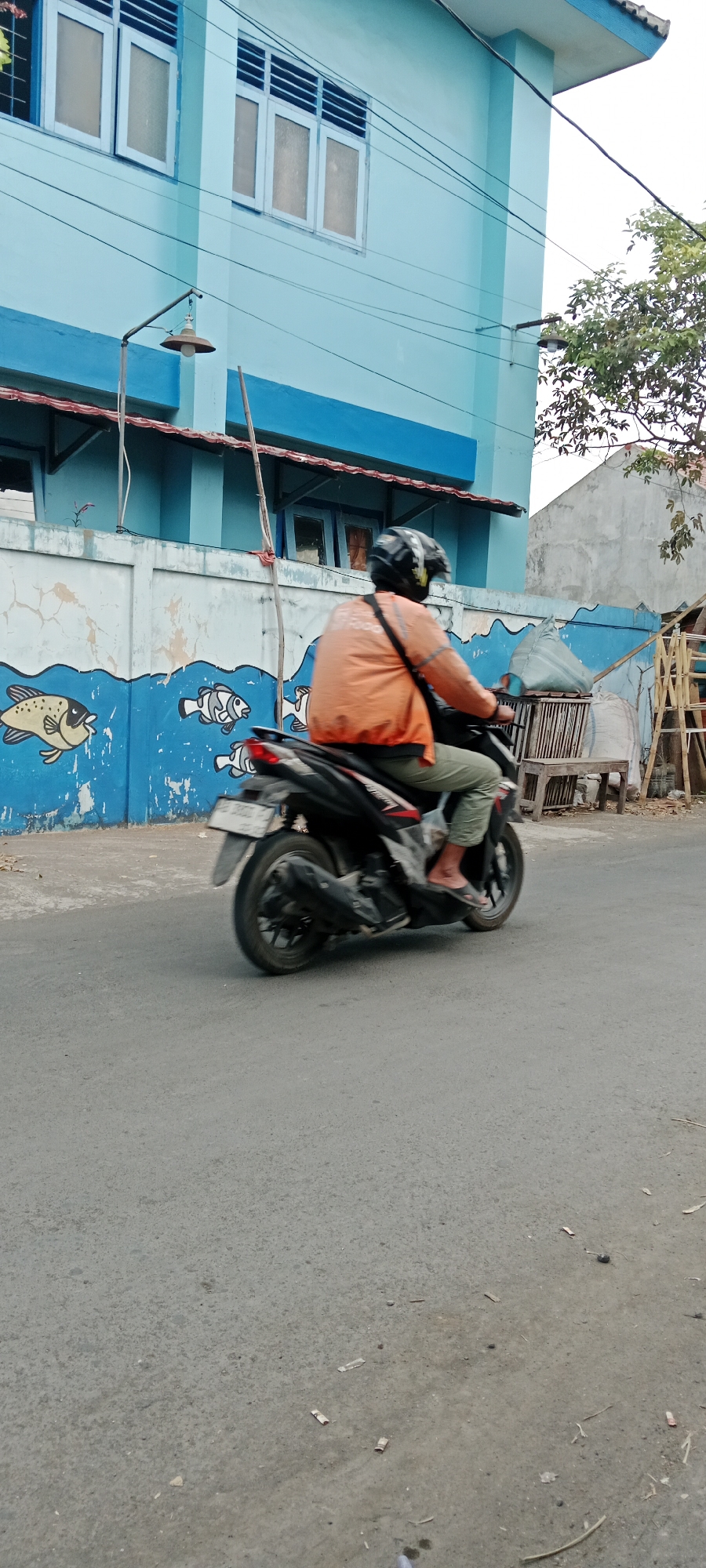 A person is riding a black motorcycle with a visible license plate that reads "AB 3833 P". The rider is wearing a black and white helmet and an orange jacket over light-colored pants and sandals. The motorcycle is in motion on a paved road. In the background, a light blue building with blue trim and white-framed windows is visible. A mural of blue waves and stylized fish is painted on a wall in front of the building. The environment appears to be a street in Purwomartani, Indonesia, with power lines running across the scene.