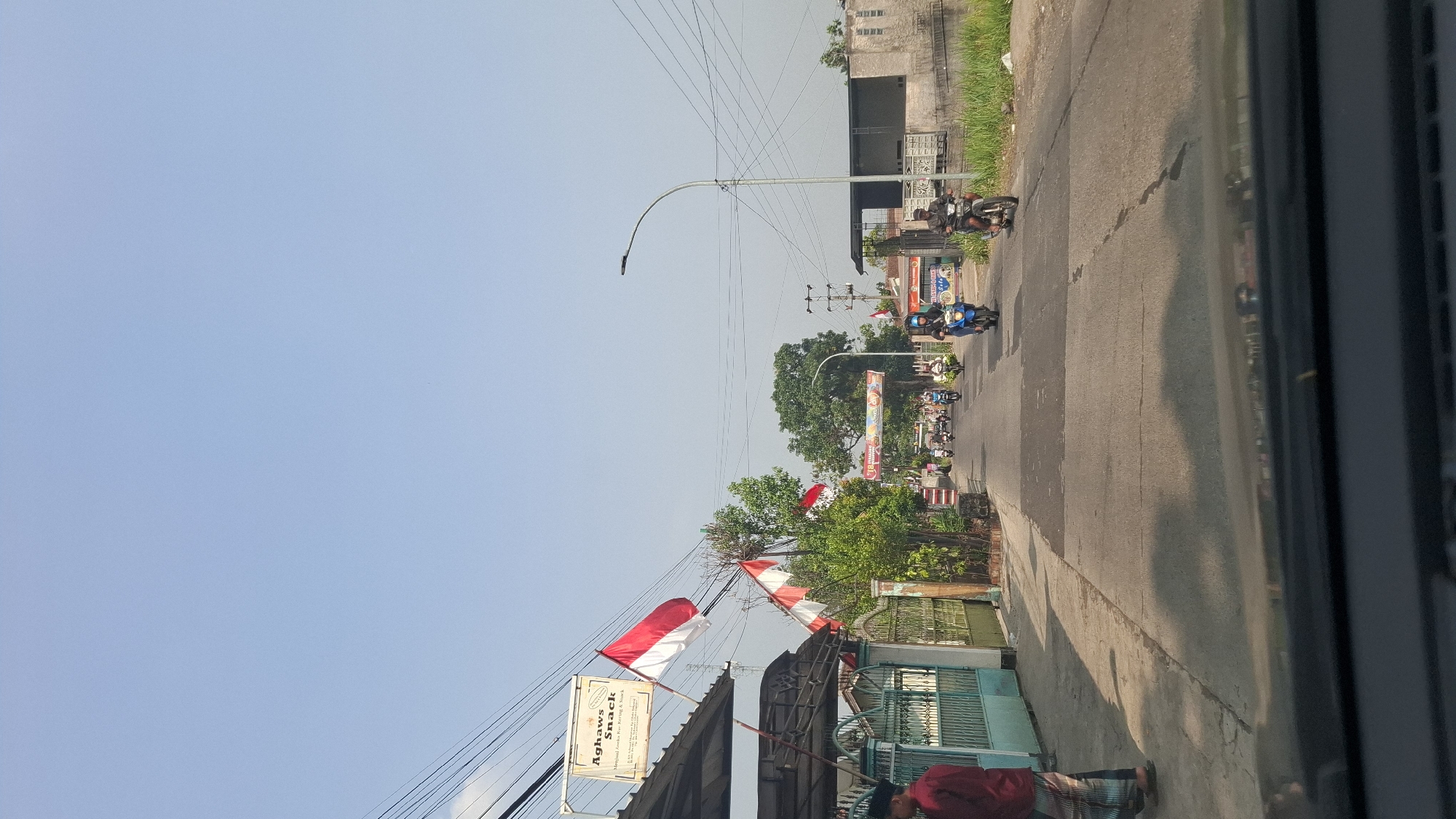 The image shows a street scene in Sukasari, Indonesia, on a clear, sunny day. The perspective is from a vehicle, looking forward down a paved road. The road has several lanes and appears to be moderately busy with motorcycles.
On the left side of the road, there is a building with a sign that reads "Aghaws Snack". Indonesian flags, red and white, are displayed on poles outside the building and along the street. A man in a red shirt and patterned pants is standing on the sidewalk to the left. Further down the street on the left, there are green gates and more trees and foliage.
On the right side of the road, there is a sidewalk with grass and some buildings. A street light with power lines is visible. Several motorcycles are riding on the road, with riders wearing helmets. One motorcycle on the right is ridden by a person in a black jacket and helmet, and another on the left is ridden by a person in a blue jacket and helmet. In the distance, there is an archway decorated with banners and more flags, suggesting a festive or celebratory occasion. The overall atmosphere is that of a typical urban street in Indonesia with some patriotic decorations.
