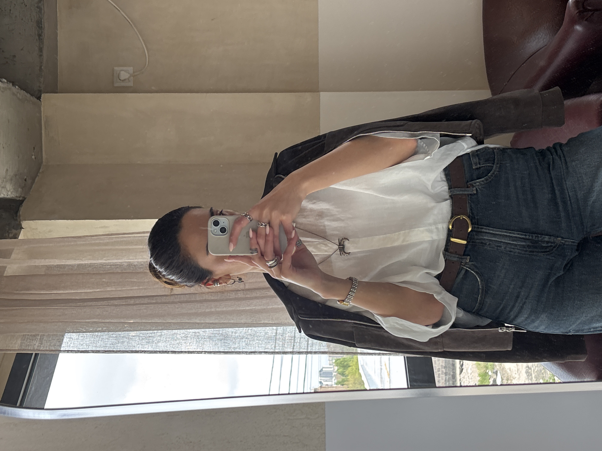The image, rotated 90 degrees counter-clockwise, presents a mirror selfie of a person, likely female, taken indoors during the daytime. The subject wears a white, loosely-fitting collared shirt, dark blue jeans, and a brown belt with a gold buckle. A dark brown or black jacket is draped over her shoulders. She is adorned with multiple silver rings on her fingers, a silver-toned watch on her left wrist, and a long necklace with a circular pendant. Her hair is pulled back, and she holds a light-colored smartphone (possibly an iPhone) with light-pink manicured nails to capture the reflection.
The indoor setting visible in the mirror features a wall with two distinct beige and light brown sections. A white power outlet and a cord are seen on the upper wall. Below, a large window, partially obscured by light, sheer curtains, reveals an outdoor urban landscape with buildings and green foliage under an overcast or cloudy sky. To the right, a dark brown piece of furniture, possibly a leather chair or sofa, is reflected. There is no visible text in the image.