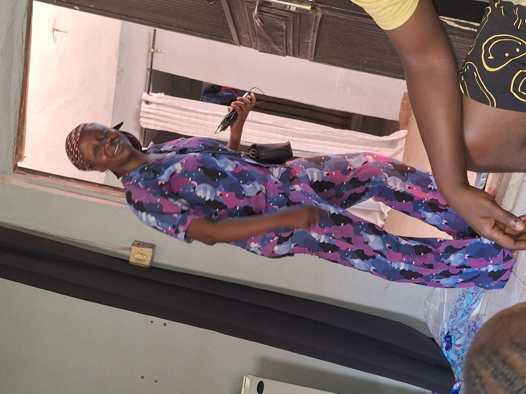 A medium shot captures a Black woman from the knees up, standing indoors and smiling at the camera. She is wearing a colorful, patterned pajama set in shades of blue, purple, and pink, and a headscarf. Her right hand holds a phone. To her left, a black curtain hangs vertically. To her right, a brown wooden door and a white wall are visible. A portion of another person's arm, wearing a yellow shirt and patterned shorts, is in the foreground on the right. The woman is positioned near an open doorway that leads to another room, where a white curtain is visible. The lighting is natural and seems to be coming from the open doorway. The overall scene appears to be a domestic interior in Sabon Tasha, Nigeria.
