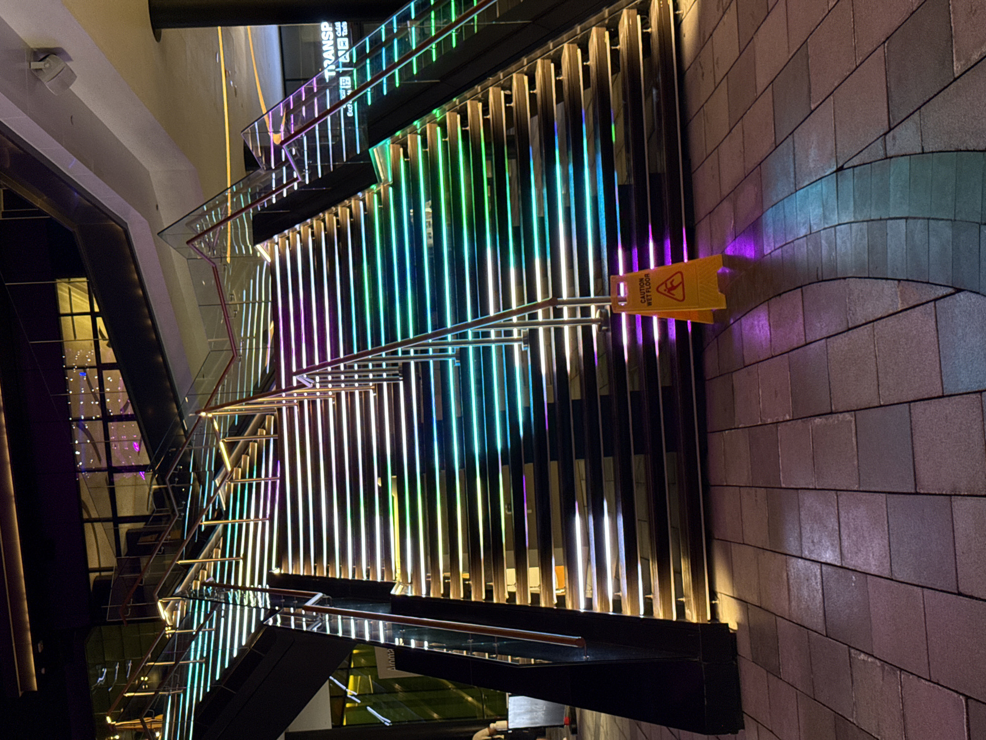 The image shows a staircase with color-changing neon lights in Dubai, United Arab Emirates. The steps are lined with vertical lights transitioning from gold, green, cyan, blue, to purple. A yellow "CAUTION WET FLOOR" sign is placed on the right side of the stairs. The walls are tiled in gray. The background includes a view of the building's interior and exterior architecture with glass windows.