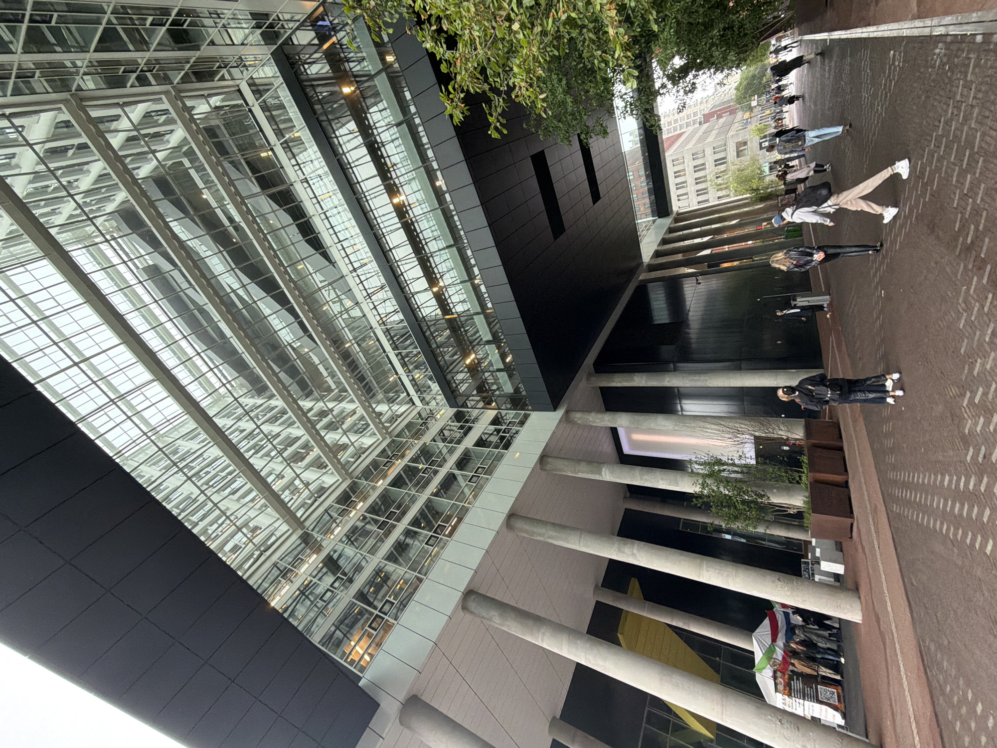 The image captures an urban scene in The Hague, Netherlands, featuring modern architecture and a public space with pedestrians. The perspective is tilted, showing a prominent multi-story building with an extensive glass and steel facade on the left, reflecting the environment. Adjacent to it, a darker, possibly black, building stands with minimalist design and limited windows.
In the foreground and extending towards the middle ground, a paved area of reddish-brown bricks stretches out, appearing somewhat damp, suggesting recent rain. Several individuals are present, walking and standing. They are casually dressed.
Beneath an overhang supported by large concrete pillars, a small gathering or event is taking place. A flag, with horizontal green, white, and red stripes, resembling the flag of Iran, is displayed prominently. Adjacent to the flag, a sign with black text on a white background and visible QR codes is set up, suggesting an informational or protest stand. A small canopy or tent structure is also part of this setup.
Green foliage from trees is visible, adding natural elements to the urban landscape. In the background, across the paved area, other multi-story city buildings with numerous windows can be seen, completing the city environment. The lighting is diffused and soft, indicating an overcast day, without direct sunlight.