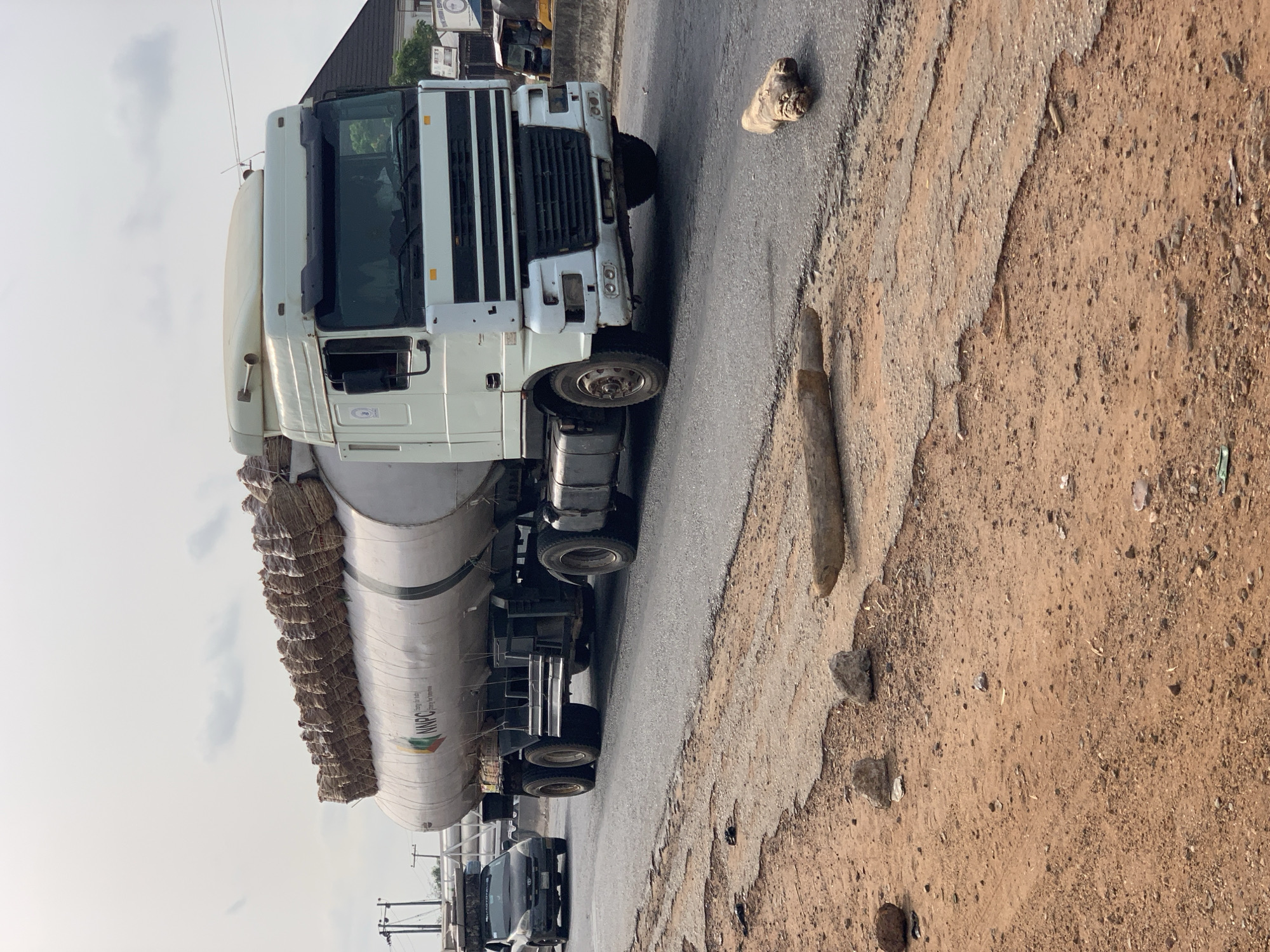 A large, white semi-truck with a silver/grey tanker trailer is shown parked or driving slowly on the shoulder of a road in Lokoja, Nigeria, under an overcast daytime sky. The truck's cab is white with a prominent black grille and bumper details.
A notable detail is the unusual cargo on top of the tanker trailer: numerous light brown, rectangular bales or bags are stacked along its entire length, secured by ropes or straps. The tanker itself features the green and white logo and text "NNPC" (Nigerian National Petroleum Corporation), along with "Energy for Life," indicating its primary function for transporting petroleum.
The road consists of a worn asphalt surface beside a wide, dry, and uneven dirt shoulder. A small, light-colored object, possibly a crumpled shoe or piece of fabric, lies on the asphalt near the truck's front wheel. In the background, other vehicles are partially visible, and distant structures are discernible beneath the cloudy sky. The scene captures a moment along a busy transport route, highlighting the multi-purpose use of commercial vehicles in the region.