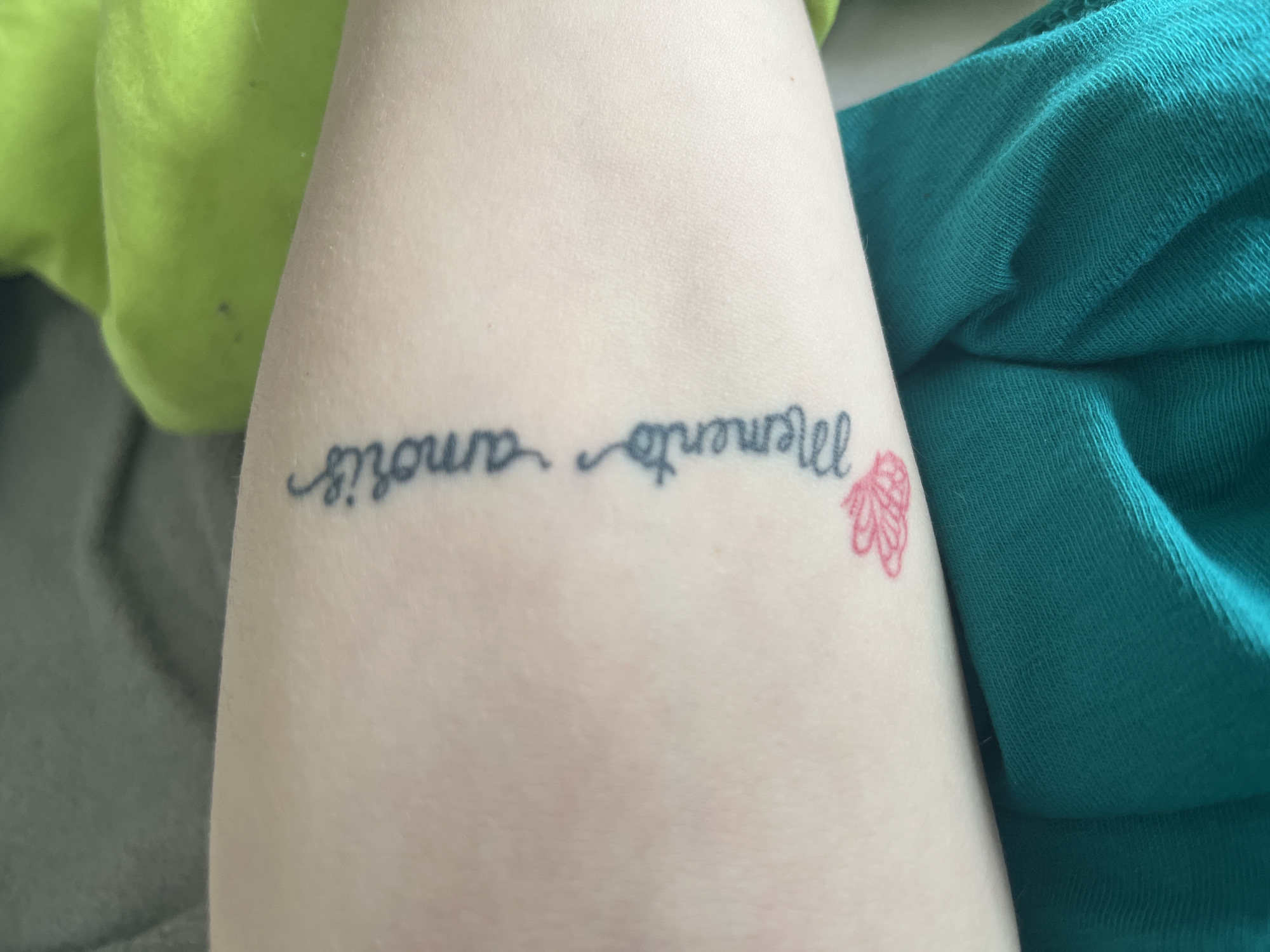 The image shows a portion of a person's arm with two tattoos. One tattoo is a black inscription reading "Memento amoris" written in cursive. To the right of the inscription is a small, pink tattoo of a floral or butterfly-like design. The arm is pale and lies on a surface with green and turquoise textiles in the background. The location is not apparent based on the information in the image and no data can be derived from the image itself. There is no evidence to determine this picture was taken in Varna, Bulgaria.