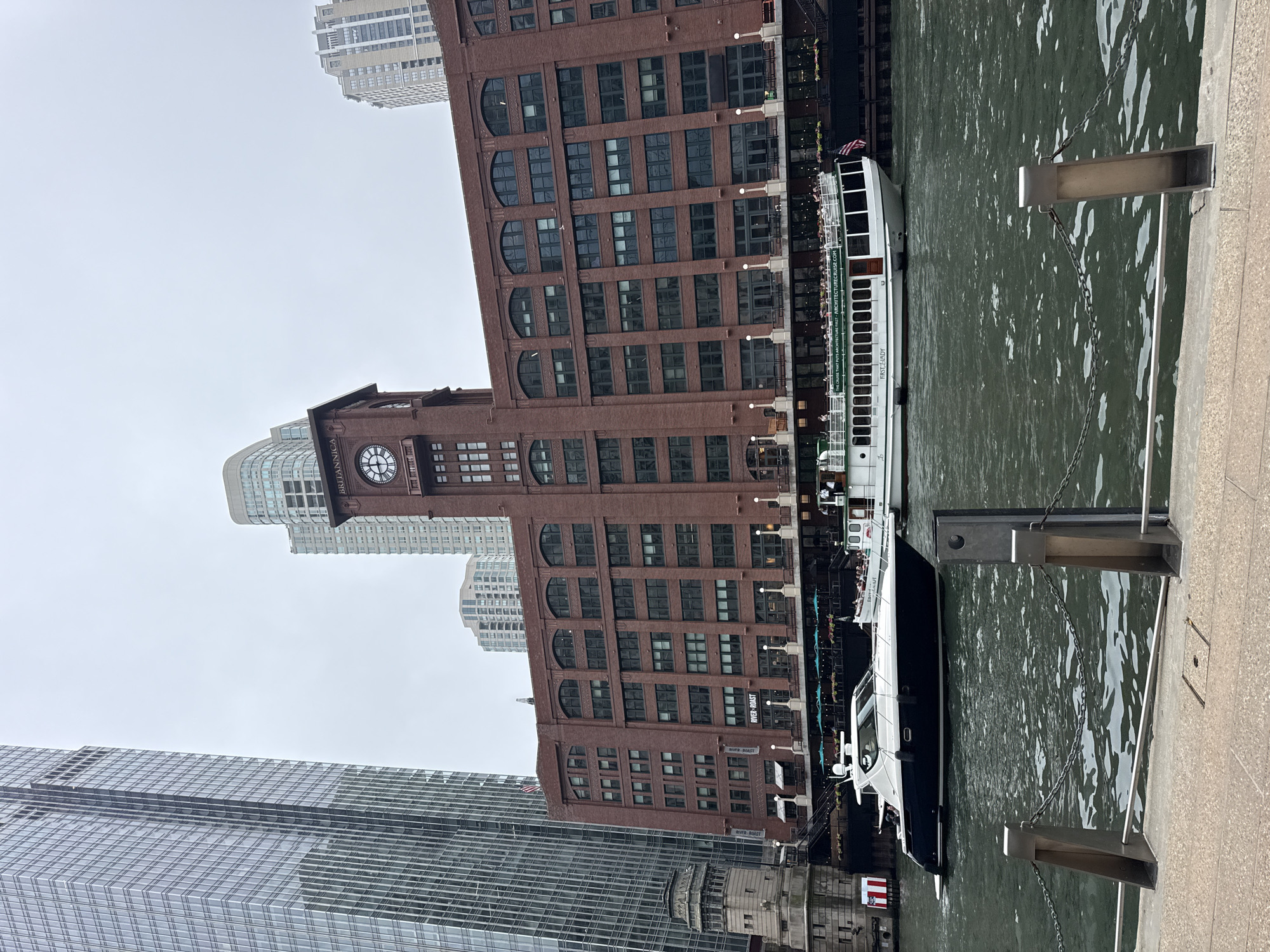 This image depicts the Chicago River in Chicago, Illinois, on a cloudy day. The scene is dominated by a large, red brick building with a prominent clock tower, identified by the sign "BRITANNICA" above the clock face. The building features numerous arched windows and appears to be a historic structure. In front of this building, a white tour boat named "FIRST LADY" is docked, with passengers visible on its upper deck. To the left of the brick building, a sleek, dark-hulled yacht is moving on the river. In the background, modern skyscrapers with glass facades rise into the overcast sky, indicating an urban environment. The foreground shows a concrete walkway and metal railings with chains, suggesting a public promenade along the river.