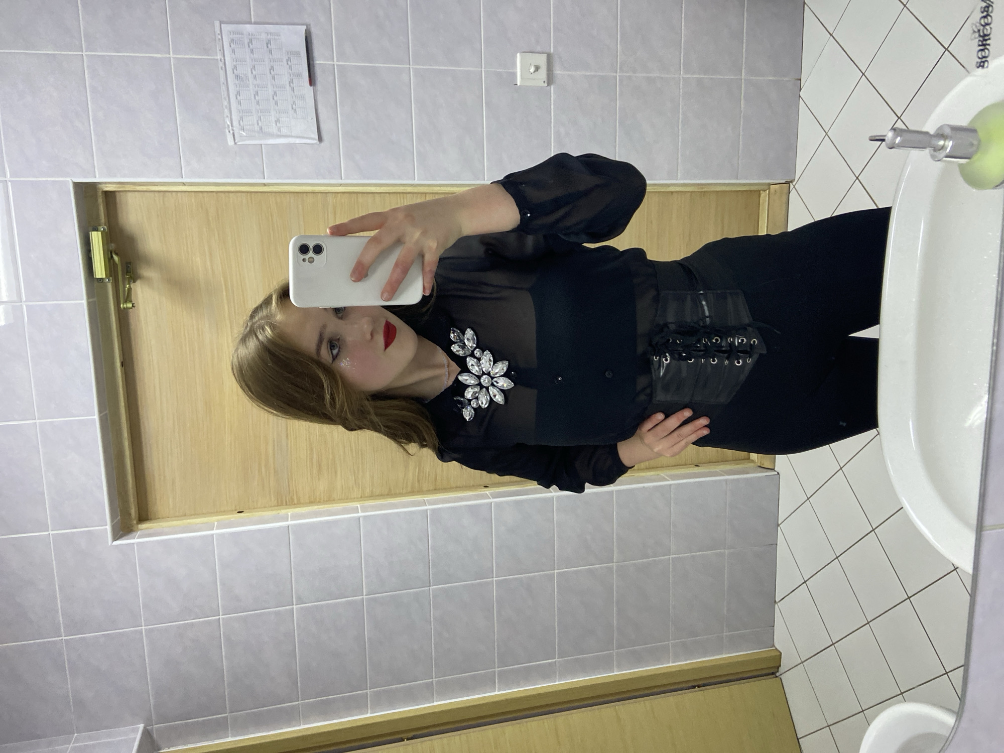 A young woman with long, wavy brown hair is taking a mirror selfie in a bathroom. She is wearing a sheer black blouse with a jeweled collar, a black bra underneath, and black pants with a black corset belt. She has red lipstick on and glitter on her eyelids. The bathroom has light grey tiled walls and white tiled floors. There is a wooden door in the background, a door closer, and a piece of paper with a grid on the wall. A soap dispenser is visible in the foreground in the reflection of the mirror. The overall impression is of a woman dressed for a night out, taking a photo in a public or semi-public restroom. The location is specified as Прикамский, Russia.