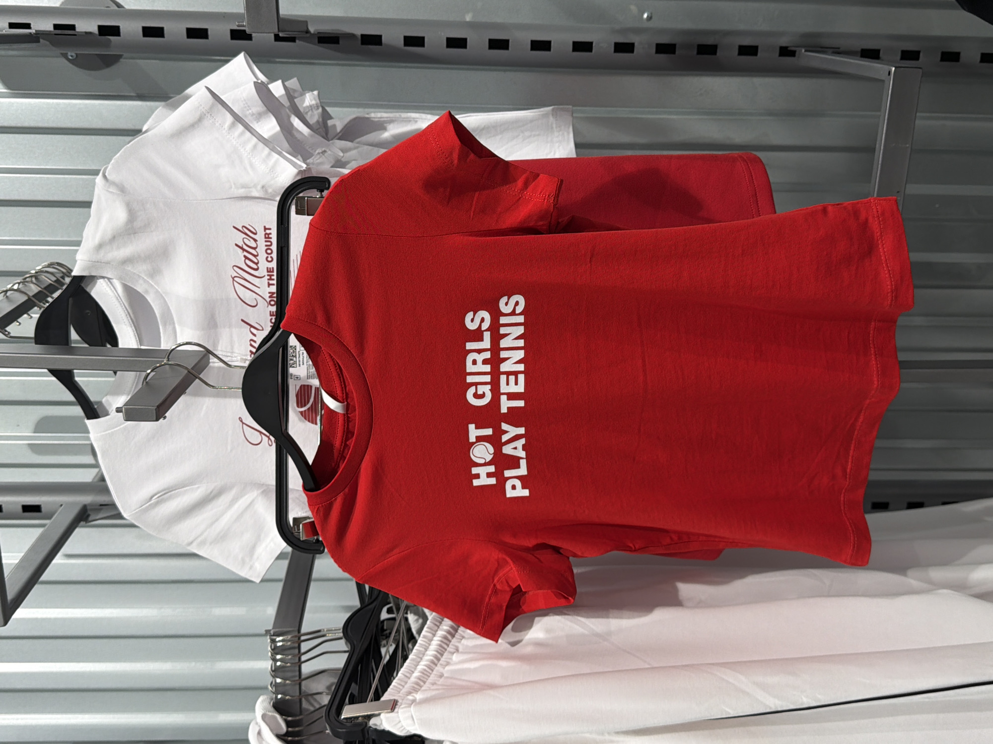 This is a medium shot, eye-level, indoor photograph taken at a retail store in Moscow, Russia. The image features several t-shirts hanging on a metal rack against a corrugated metal wall. In the foreground, a bright red t-shirt is prominently displayed, with white text that reads "HOT GIRLS PLAY TENNIS." A graphic of a tennis ball is integrated into the text. Behind the red t-shirt, a white t-shirt is partially visible, displaying a script-style text that appears to say "Grand Match" and below that, "ON THE COURT." Further to the left, a pair of white sweatpants are also hanging on the rack. The lighting is even and appears to be artificial. There are no people or discernible animals present in the image.