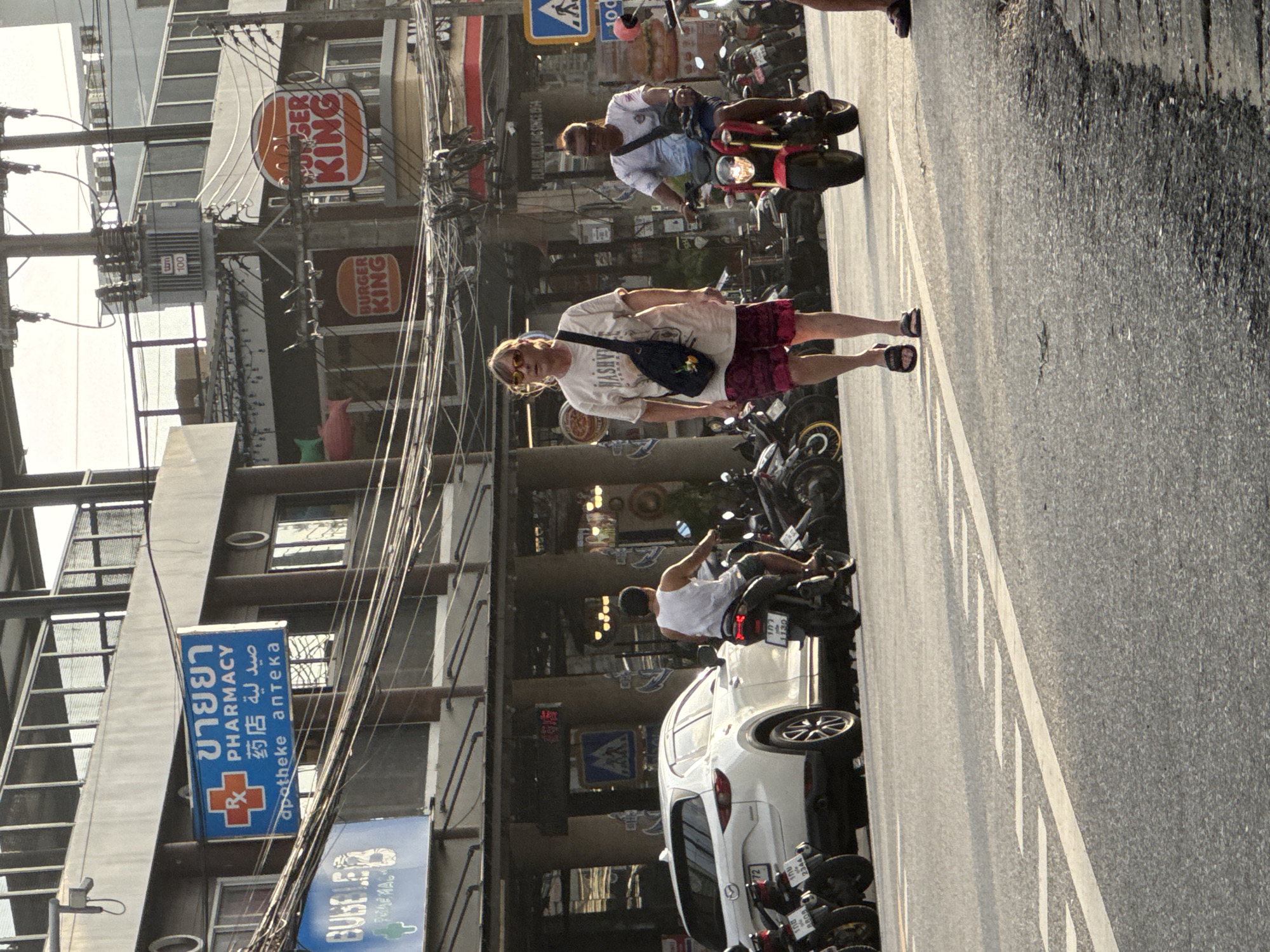 The image depicts a street scene in Karon, Thailand, likely in the late afternoon given the lighting. In the foreground, a woman wearing sunglasses, a light-colored t-shirt, and patterned shorts walks on the right side of the road, carrying a dark crossbody bag. To her right, a man rides a red motorcycle, also wearing sunglasses and a t-shirt. Behind them, a white SUV is parked on the left side of the road, with several motorcycles parked in a row further down.
The background features commercial buildings with visible signage, including a "Pharmacy" sign with Thai and English text, and several "Burger King" signs. Overhead, a complex network of electrical wires is prominent against the sky. The street is paved and appears to be a typical urban thoroughfare with painted road markings. Several other individuals and motorcycles are visible further in the background, suggesting a moderately busy street. The overall environment is consistent with a commercial area in a Southeast Asian city.
