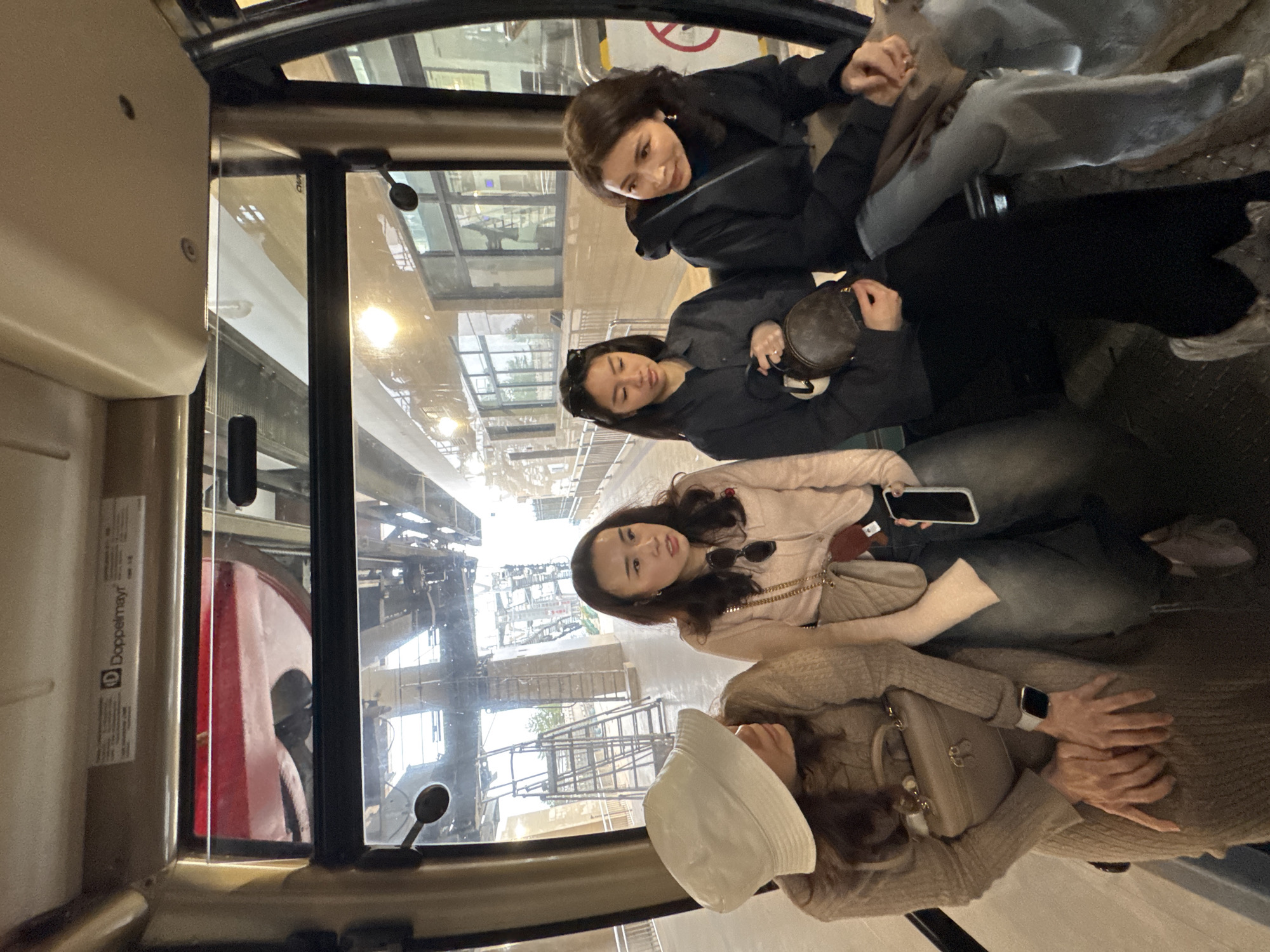 The scene takes place inside a Doppelmayr cable car cabin at a station in Wulingyuan, China, during daylight. Four adult East Asian females are seated across the cabin.
From left to right:
1. A female in a light-colored bucket hat and a ribbed, beige long-sleeved dress or top. She holds a small, light-colored handbag and wears a smartwatch on her left wrist. Her gaze is directed to her right.
2. A female with long dark hair, wearing a light pink collared shirt and dark pants, with sunglasses hanging from her shirt. She holds a dark smartphone with a light case and has a small bag with a chain strap across her body. Her mouth is slightly open, and she looks slightly to her left.
3. A female with long dark hair, wearing sunglasses atop her head, a dark long-sleeved top, and dark pants. She holds a small, dark, rounded handbag in her lap. She is looking downwards. A ring is visible on her left ring finger.
4. A female with brown hair, wearing a dark hooded jacket over a dark turtleneck and light blue jeans. A brown bag rests on her lap. She is looking to her left with a slight smile.
Through the cabin's large windows, elements of the cable car station infrastructure are visible, including metal girders, support beams, and a building with multiple glass windows. A tall, multi-rung metal ladder or scaffolding structure is present outside to the left. The cabin appears to be stationary at a platform.