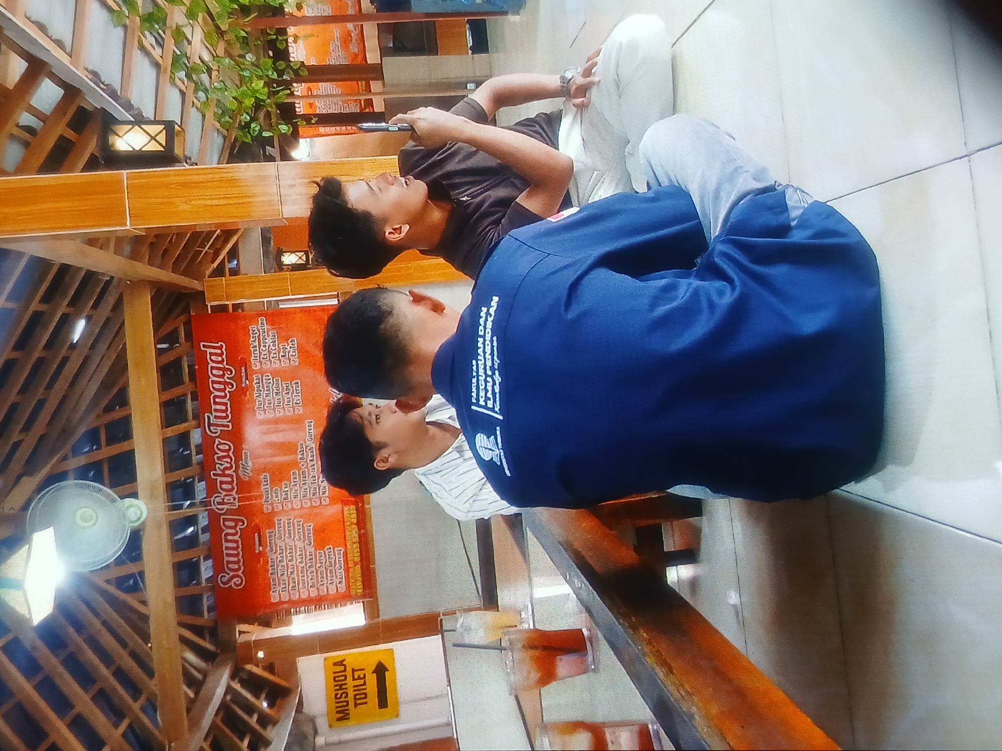 A medium shot shows three young men sitting on a tiled floor in an outdoor seating area, possibly a food stall or restaurant, with a thatched roof supported by wooden beams. In the background, a large red banner with Indonesian text, "Saung Bakso Tunggal," is visible, listing food and drink items. To the left, a yellow sign with a black arrow points to "MUSHOLLA TOILET."
Two men are in the foreground, viewed from the back. One wears a navy blue polo shirt with white lettering, the other a dark brown polo shirt. The man in the brown shirt holds a smartphone in his right hand, looking towards the left of the frame and appearing to be speaking or gesturing. A third man, wearing a white and pinstripe shirt, sits to the left of the two in the foreground, facing towards the right and looking at the man in the brown shirt.
On a wooden table in the middle ground, two glasses with iced drinks are visible. The environment appears to be a casual, open-air dining space. The overall lighting suggests daytime.
