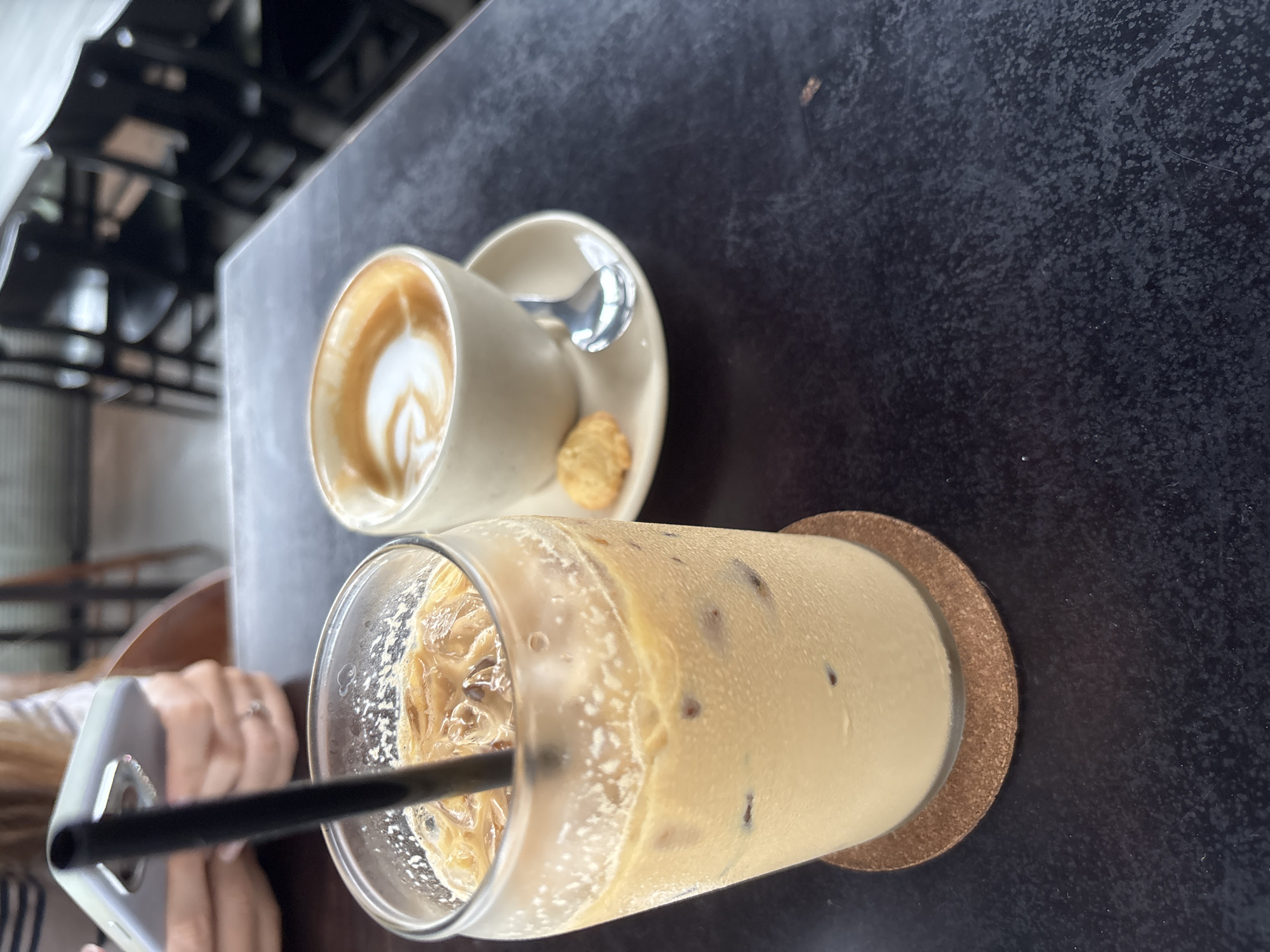 The photograph presents a top-down view of a table setting with two beverages, likely taken in Kuta Selatan, Indonesia. On the left is a small ceramic cup of latte with visible latte art, resting on a matching saucer. A small cookie sits on the saucer next to the cup. A silver spoon is also on the saucer. To the right is a tall glass filled with ice and a light brown liquid, possibly iced coffee or tea. A black straw is inserted in the glass, which sits on a round cork coaster. The table is dark and appears to be made of a material with a coarse texture. A hand is visible holding a phone, and architectural structures are visible in the background.