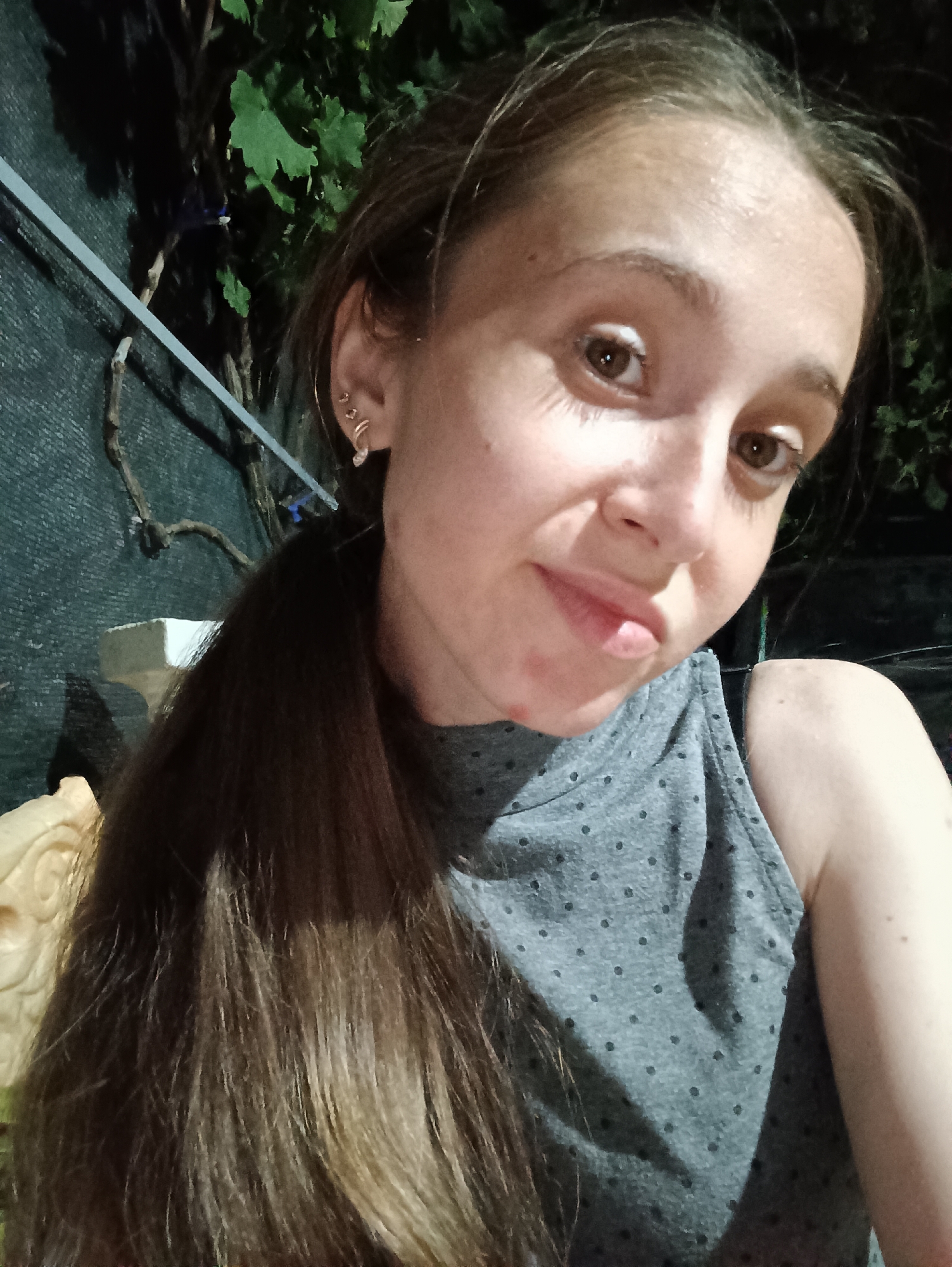 The image shows a close-up, front-facing selfie of a young Caucasian woman with long, dark brown hair tied in a ponytail. She is wearing a gray sleeveless top with a polka dot pattern. Her facial features include brown eyes, a slightly parted mouth, and visible eyelashes. There are some blemishes on her face. She is also wearing multiple earrings in her left ear. The background appears to be outdoors at night, with some foliage and a dark, possibly netted, structure behind her. A beige, carved object is partially visible to the left of the frame. The lighting suggests it is artificial and positioned to illuminate her face. No specific location in Vypasne, Ukraine can be determined from the image itself.