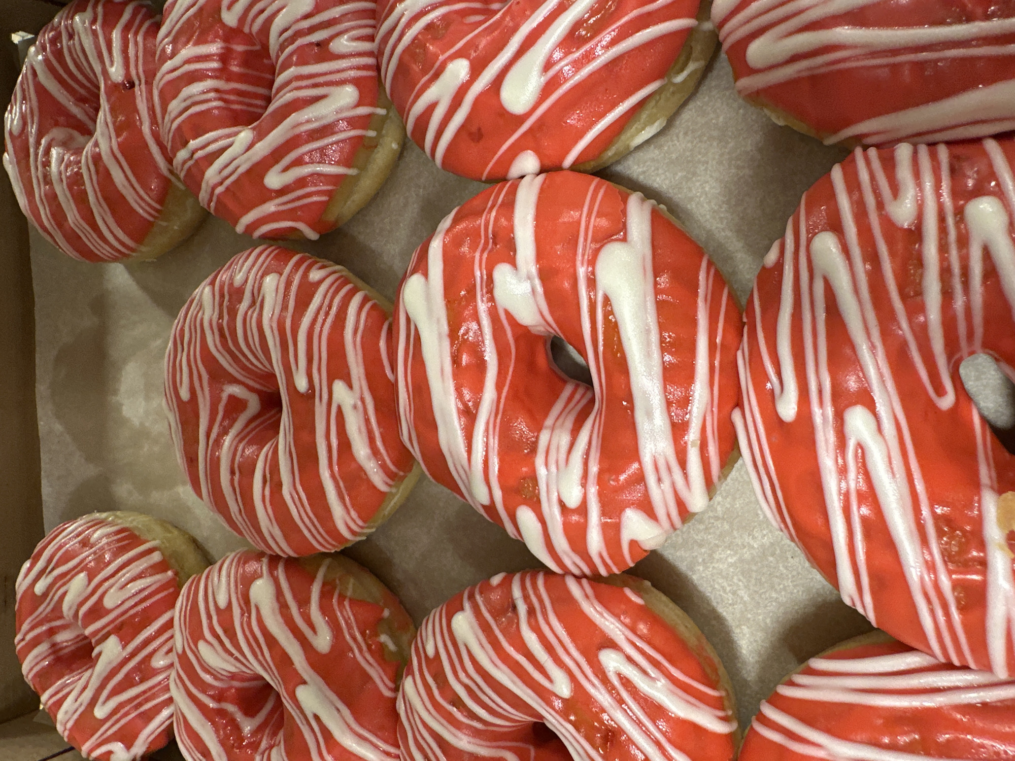 A close-up, overhead shot depicts a box of a dozen donuts. The donuts are ring-shaped and topped with a vibrant red glaze. A white drizzle decorates the red glaze in a linear pattern. The donuts are arranged closely together in the box, filling the space. The location is not discernible from the image.