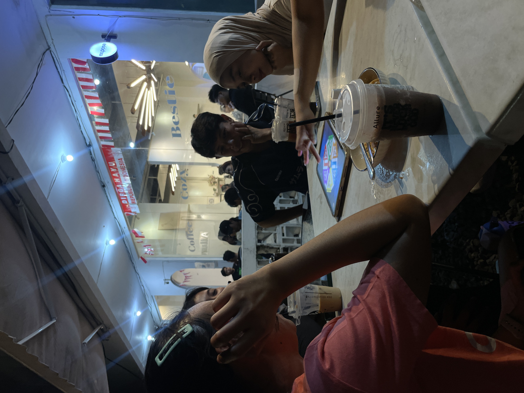 This image depicts a group of young adults gathered around a table at a cafe, likely in Manado, Indonesia, indicated by the Indonesian flag colors visible in the background signage and the cafe's name "Bestie" which is a common term used in Indonesia. The scene is set at night, with artificial lighting illuminating the outdoor seating area.
In the foreground, a person wearing a pink t-shirt is partially visible, with their left arm resting on the table. To their right, a tablet computer is laid out on the table, displaying a colorful game interface. A young woman wearing a hijab is looking intently at the tablet, her hands resting on the table. Next to her, a young man in a black t-shirt is also observing the tablet, with his chin resting on his hand. Several drinks, including a chocolate beverage in a clear cup with the brand "Allure" visible, are also on the table.
In the background, through the glass facade of the cafe, other patrons are visible. The cafe interior appears modern and well-lit, with stylish light fixtures. The signage above the entrance features the Indonesian flag colors (red and white stripes) and the words "DIRGAHAYU REPUBLIK INDONESIA," suggesting a celebration or national holiday. Another sign on the building reads "expose," likely the name of the establishment or a featured event. The overall atmosphere suggests a casual social gathering at a popular hangout spot.