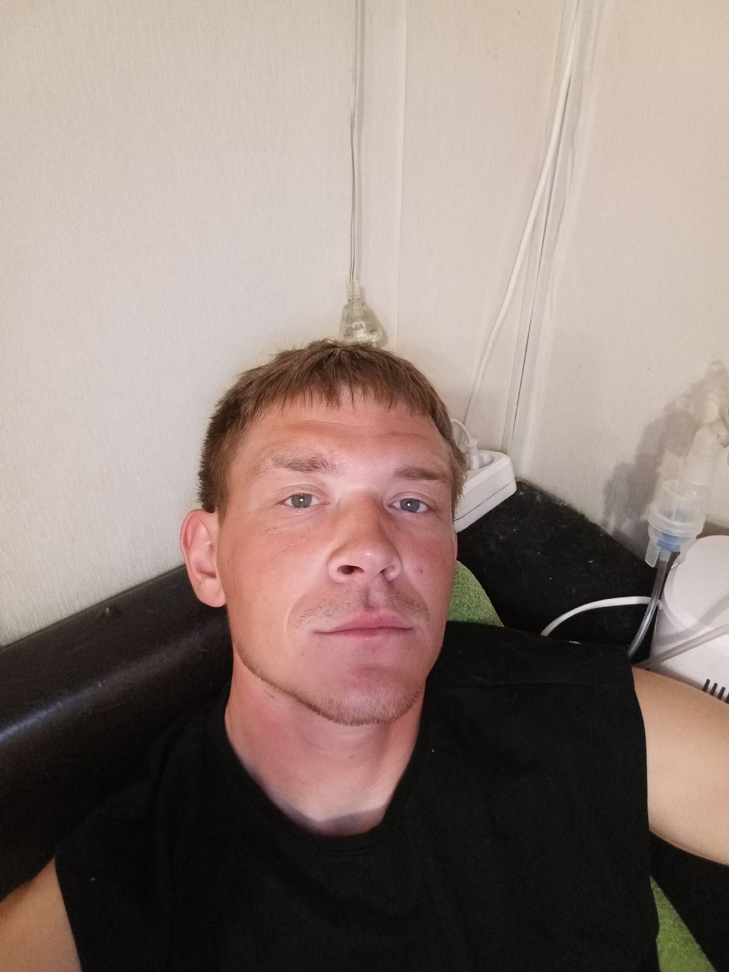 An adult male is depicted in a close-up, selfie-style photograph, positioned from the chest upwards. The individual has a light complexion, blue eyes, short light brown hair with a fringe, a faint mustache, and a short beard along his jawline. A reddish tint is noticeable on his neck. He wears a plain black sleeveless top and maintains a neutral expression while looking directly at the camera. The background consists of a dark, possibly padded surface to the left, and an off-white wall with subtle vertical textures to the right. On a dark surface to the right, a white medical device, identifiable as a nebulizer with attached clear tubing and a clear medication cup, is visible. Above this device, a white electrical outlet with a white cord plugged in is mounted on the wall, alongside other white cords running vertically. A green fabric object is partially visible between the male and the medical equipment. The precise location within the city of Chaikovsky, Russia, is not discernible from the image content.