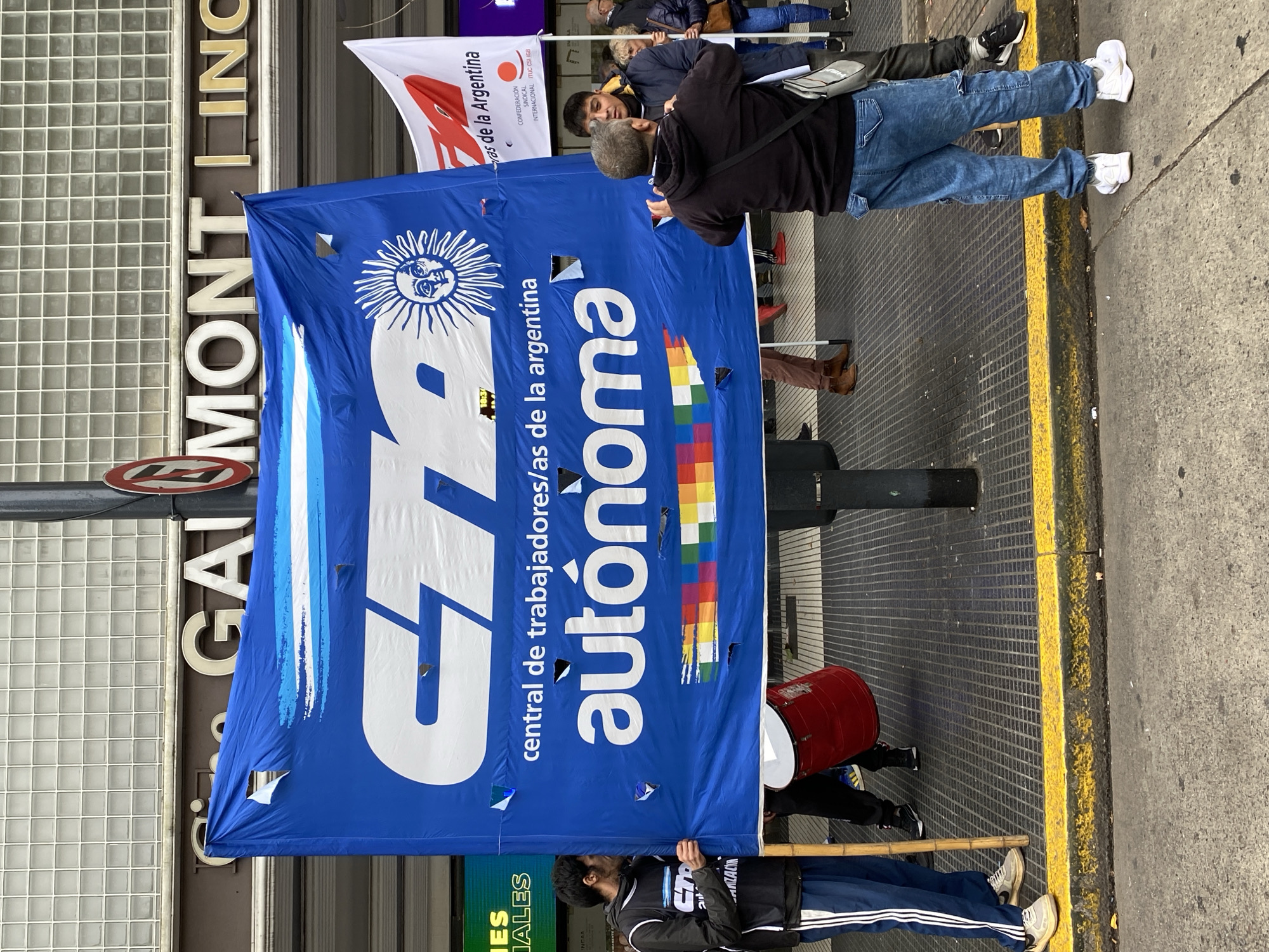The image depicts a street scene in Buenos Aires, Argentina. A large, blue banner with the acronym "CTA" (Central de Trabajadores/as de la Argentina) is prominently displayed. The banner also features the word "autónoma" (autonomous), a sun symbol, and a design with rainbow colors. The banner has some tears and holes.
Behind the banner, a sign reads "GAI MONTLING". Several people stand around the banner, including one man appearing to take a photo of it. A drum is also visible. A smaller flag, bearing the words "ras de la Argentina" and a logo, is partially visible above the blue banner.
The setting appears to be an urban environment, with a building facade and sidewalk visible. The presence of the CTA banner indicates a gathering or demonstration related to workers' rights or similar social issues.