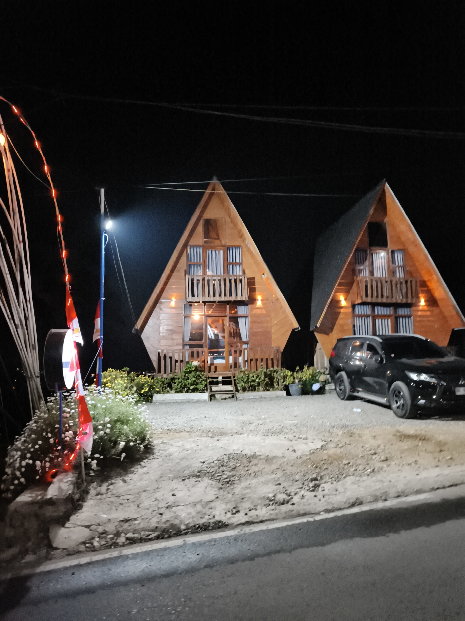 The image shows two A-frame wooden cabins at night in Dieng Kulon, Indonesia. The cabins have multiple windows and balconies, with lights visible inside and outside. A black SUV is parked in front of the right cabin. To the left of the cabins, there are bamboo poles adorned with red string lights and two Indonesian flags. A patch of white flowers is visible in the foreground, and the ground is covered with gravel and dirt. The scene appears to be a roadside accommodation or tourist spot.